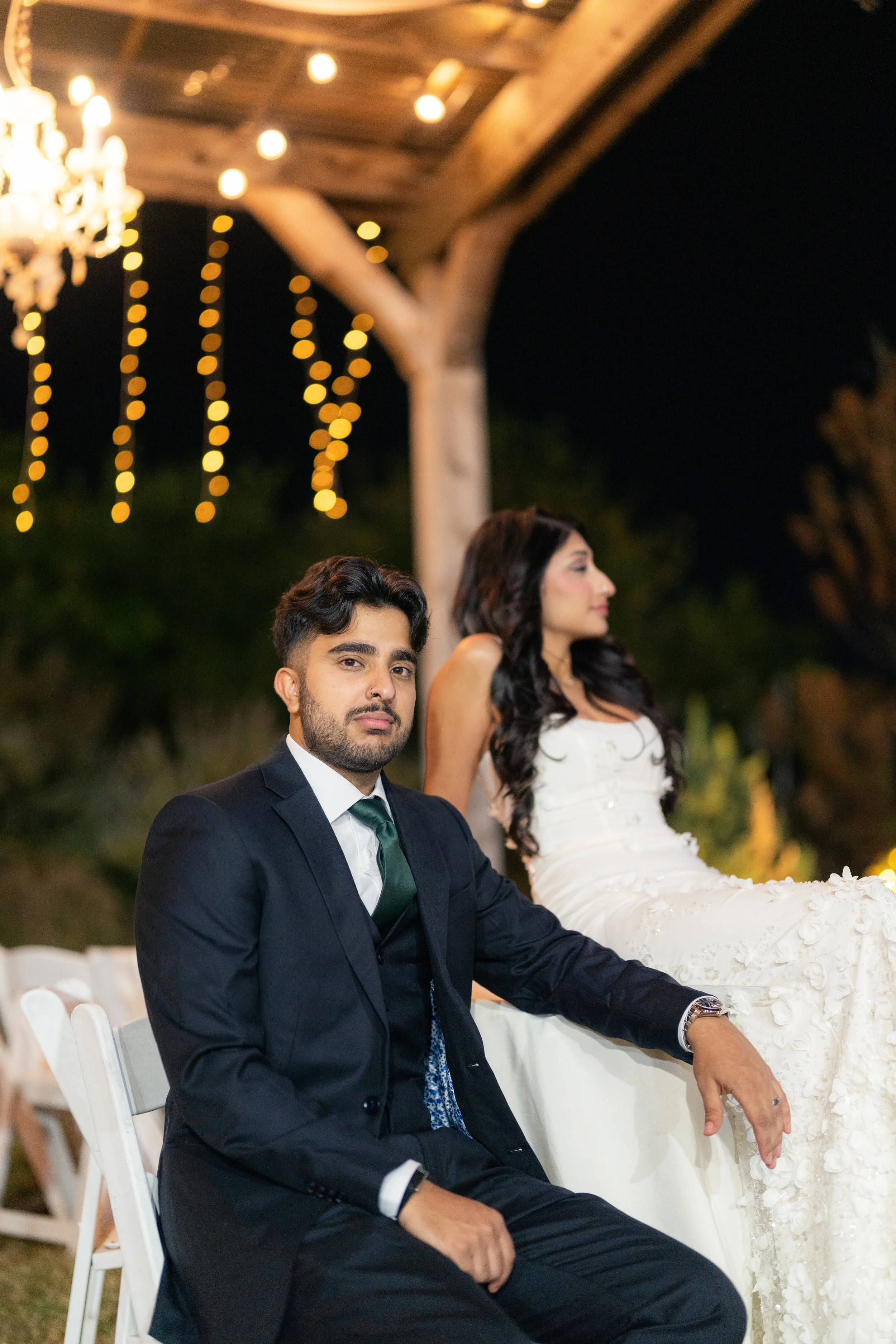 A man in a black suit and a woman in a white dress sitting at a nighttime outdoor event with string lights and a chandelier overhead.