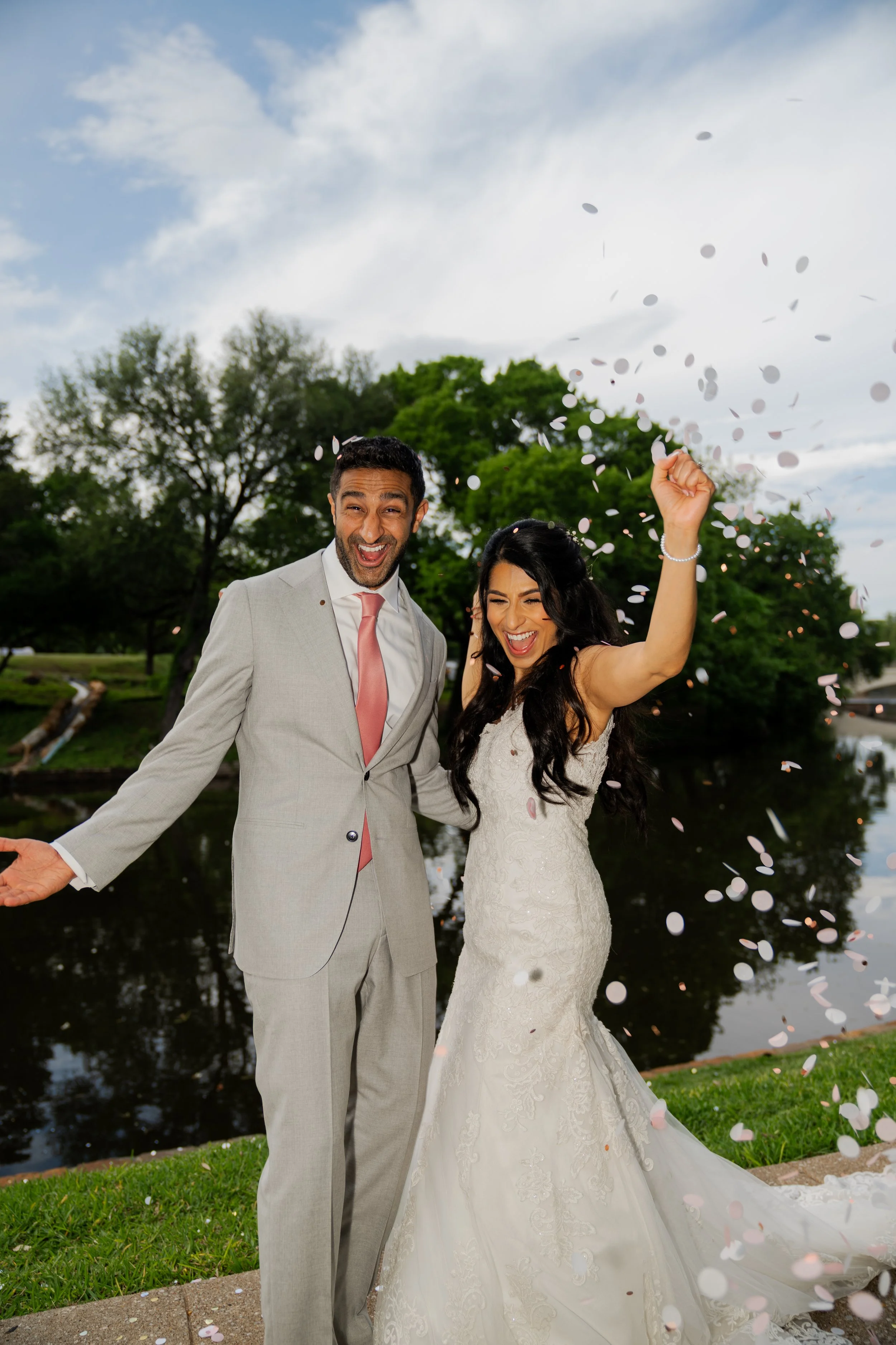 A newly married couple celebrating outdoors with confetti falling around them by a pond, surrounded by trees and a cloudy sky.