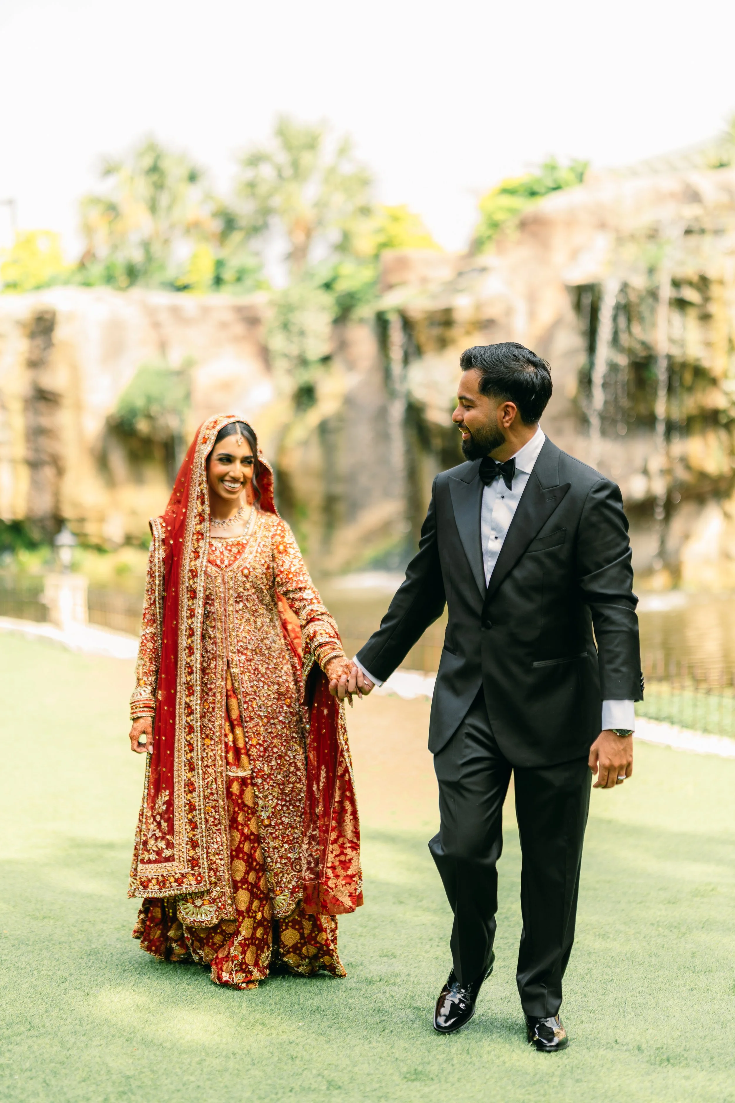 A bride and groom walking hand in hand outdoors on grass, smiling at each other. The bride is dressed in a traditional red and gold embroidered bridal outfit with a red veil, and the groom is wearing a black tuxedo with a bowtie.