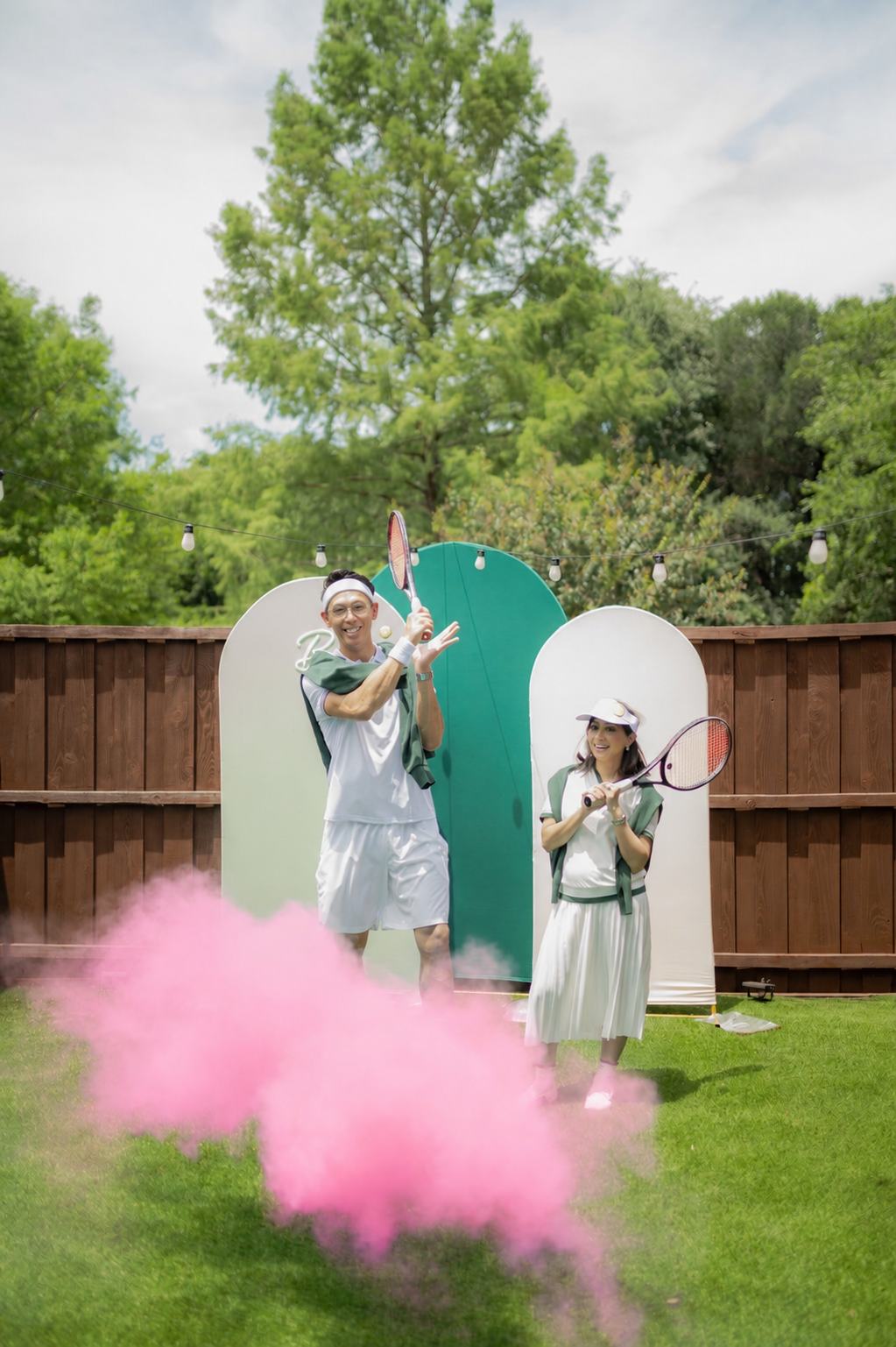 Two people in tennis attire holding tennis rackets in a backyard with a wooden fence, green grass, and trees in the background. Pink smoke on the ground, outdoor string lights overhead, and decorative panels behind them.