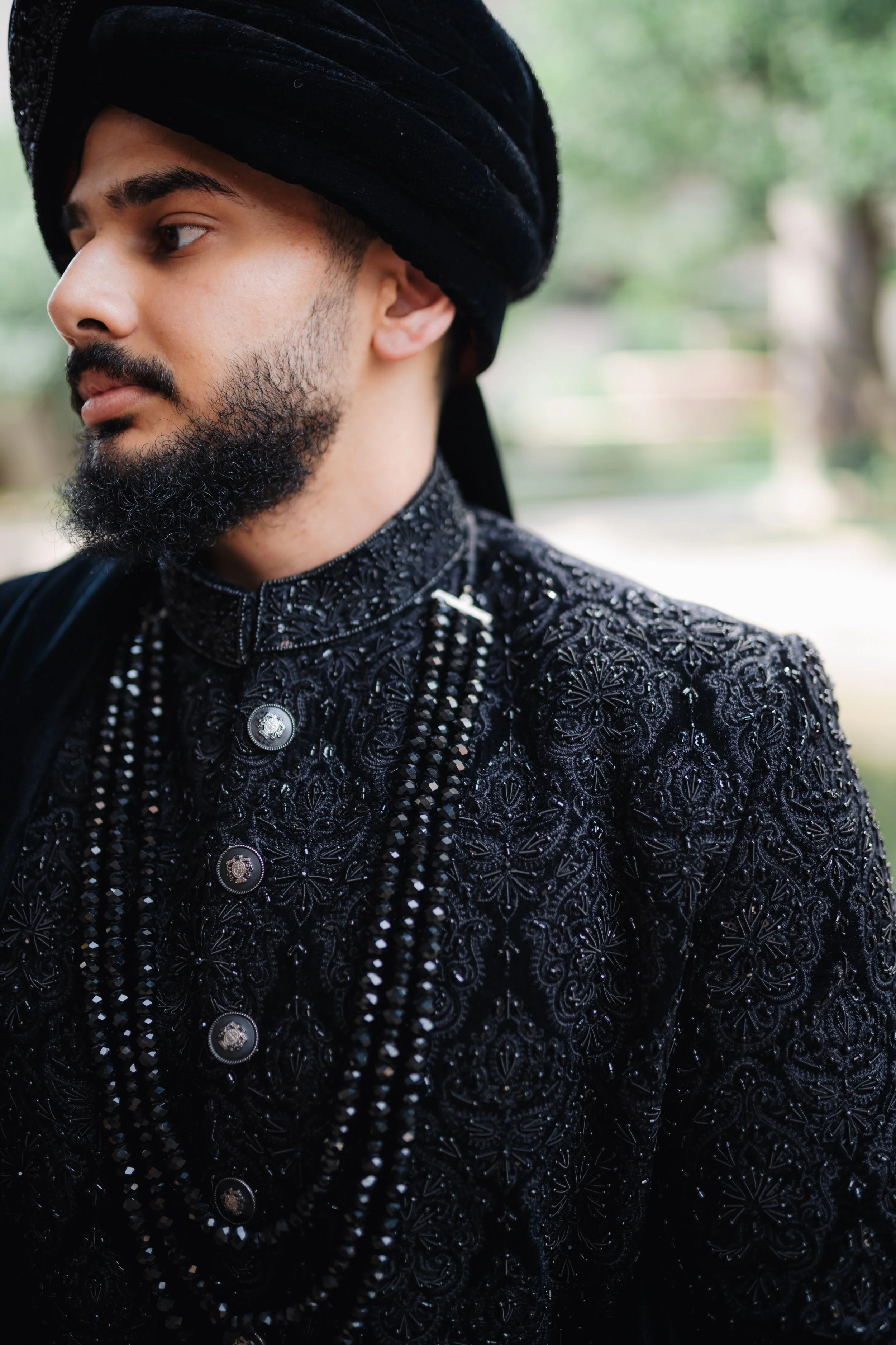 Close-up of a man in traditional black Indian attire with intricate embroidery and silver buttons, wearing a black turban, standing outdoors with greenery in the background.