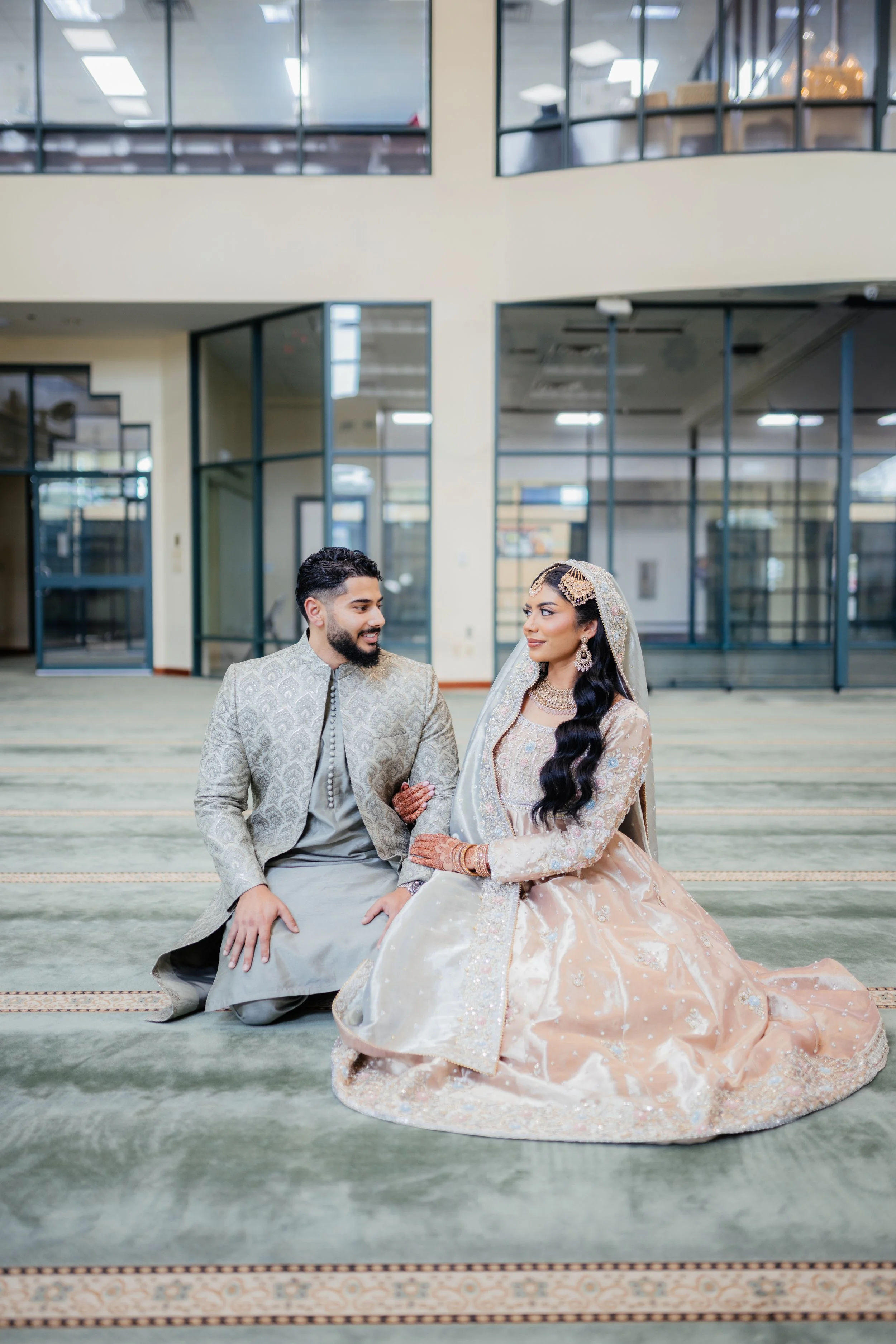 A bride and groom dressed in traditional South Asian wedding attire, sitting on the carpeted floor in a spacious, modern indoor venue with large glass windows.