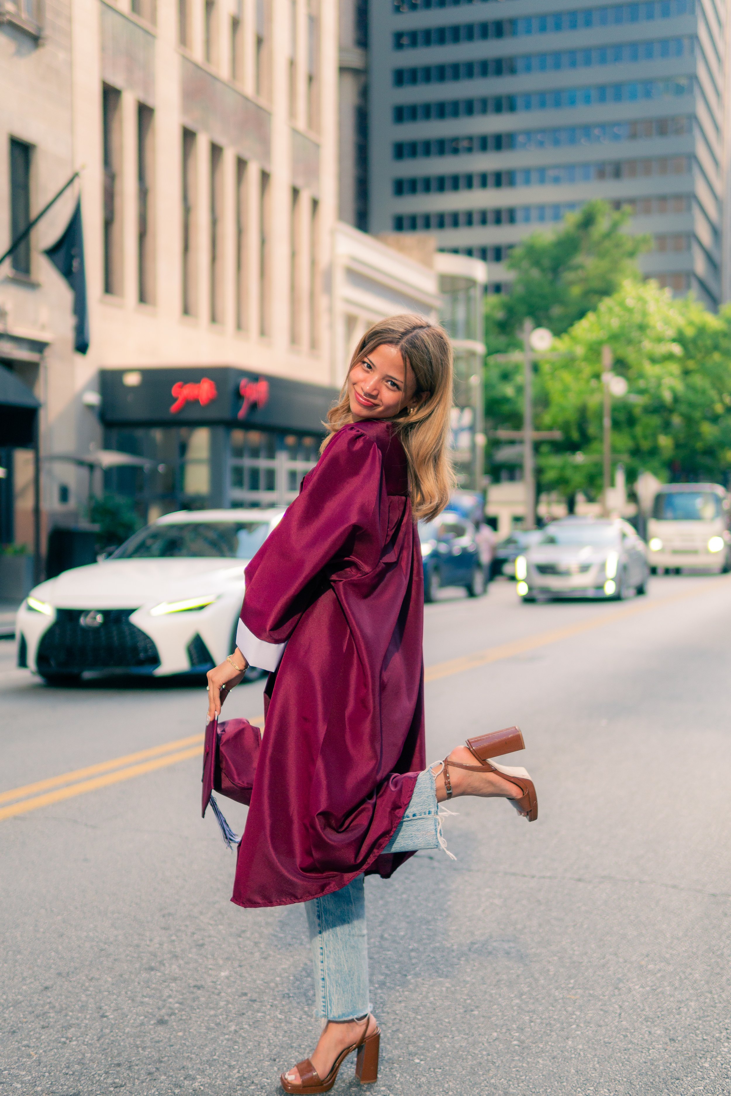 A young woman in a burgundy graduation gown is smiling and playfully lifting her leg while walking on a city street. She holds her cap in one hand and wears high-heeled shoes, with tall buildings, cars, and green trees in the background.