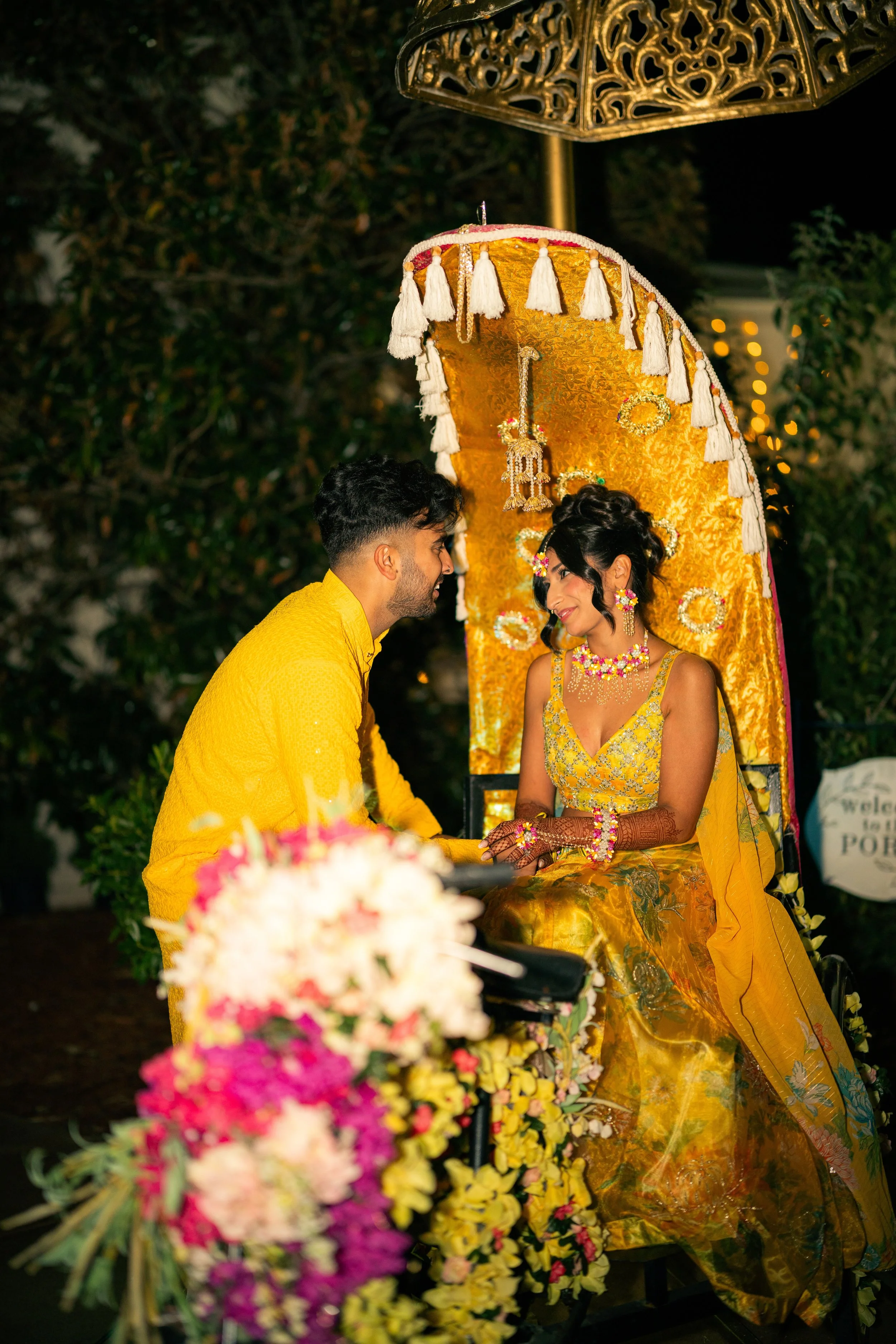A couple at a traditional Indian wedding ceremony, dressed in yellow, sitting under a decorated yellow canopy with floral arrangements in the foreground.