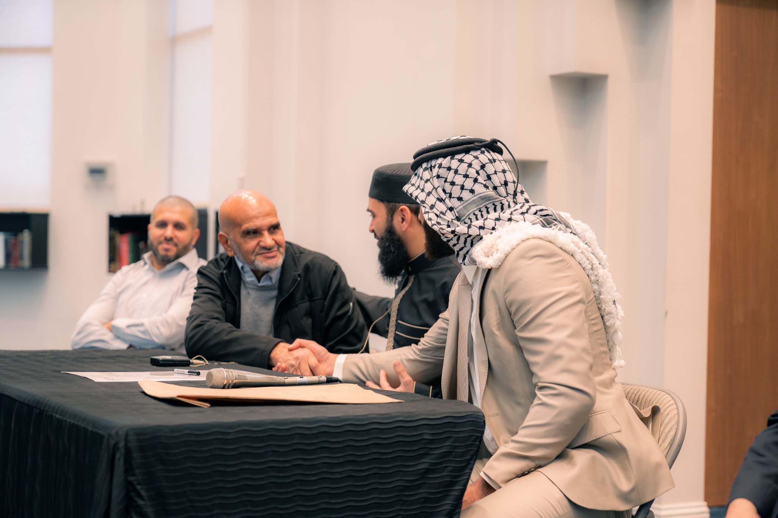 Group of four men, two of Middle Eastern descent and two of South Asian descent, shaking hands in a professional setting, with others observing, at a table with microphones and papers.
