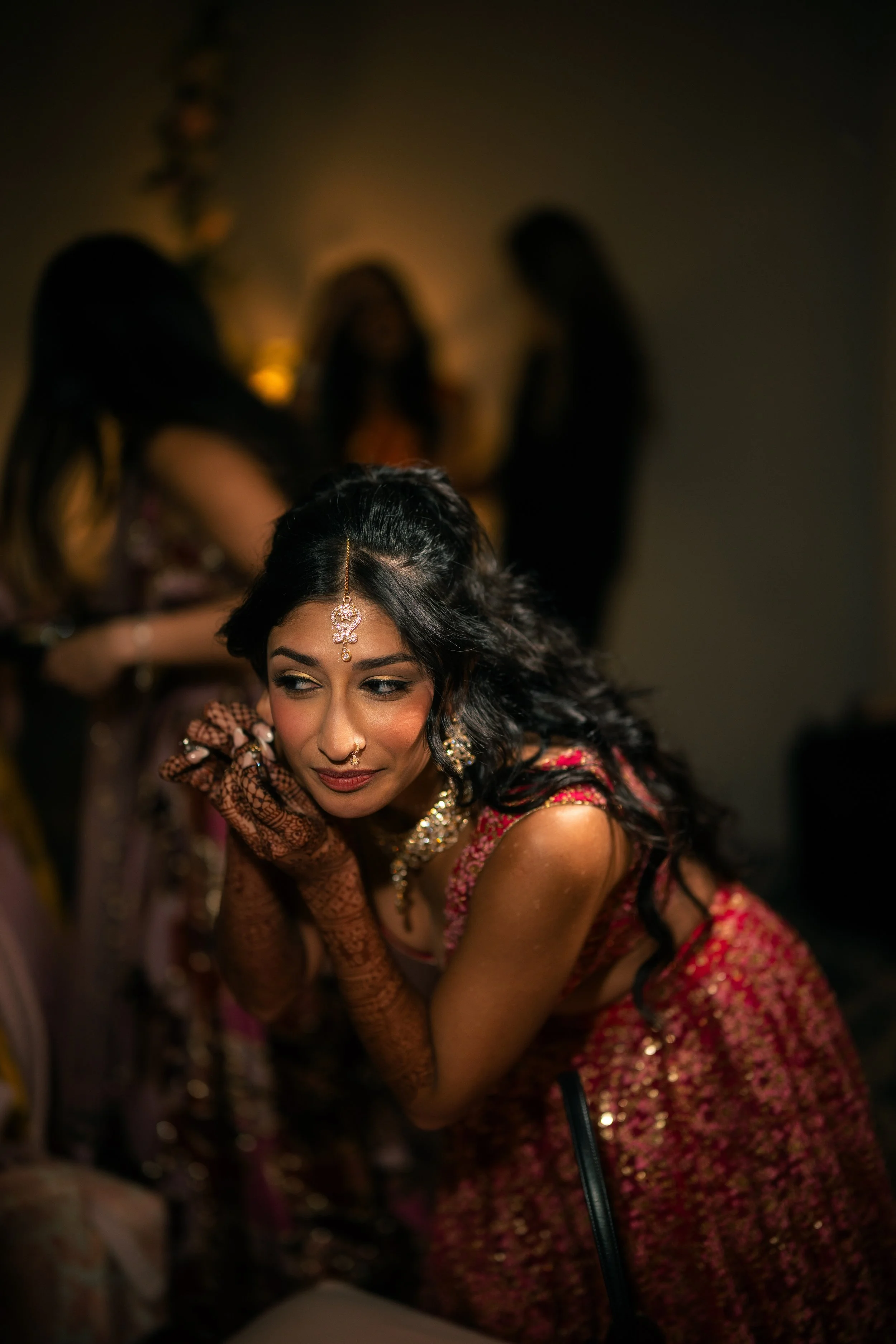 A woman in traditional Indian attire with jewelry and henna tattoos, leaning forward with hands folded near her face, at what appears to be a cultural or ceremonial event, with three other women in the background.