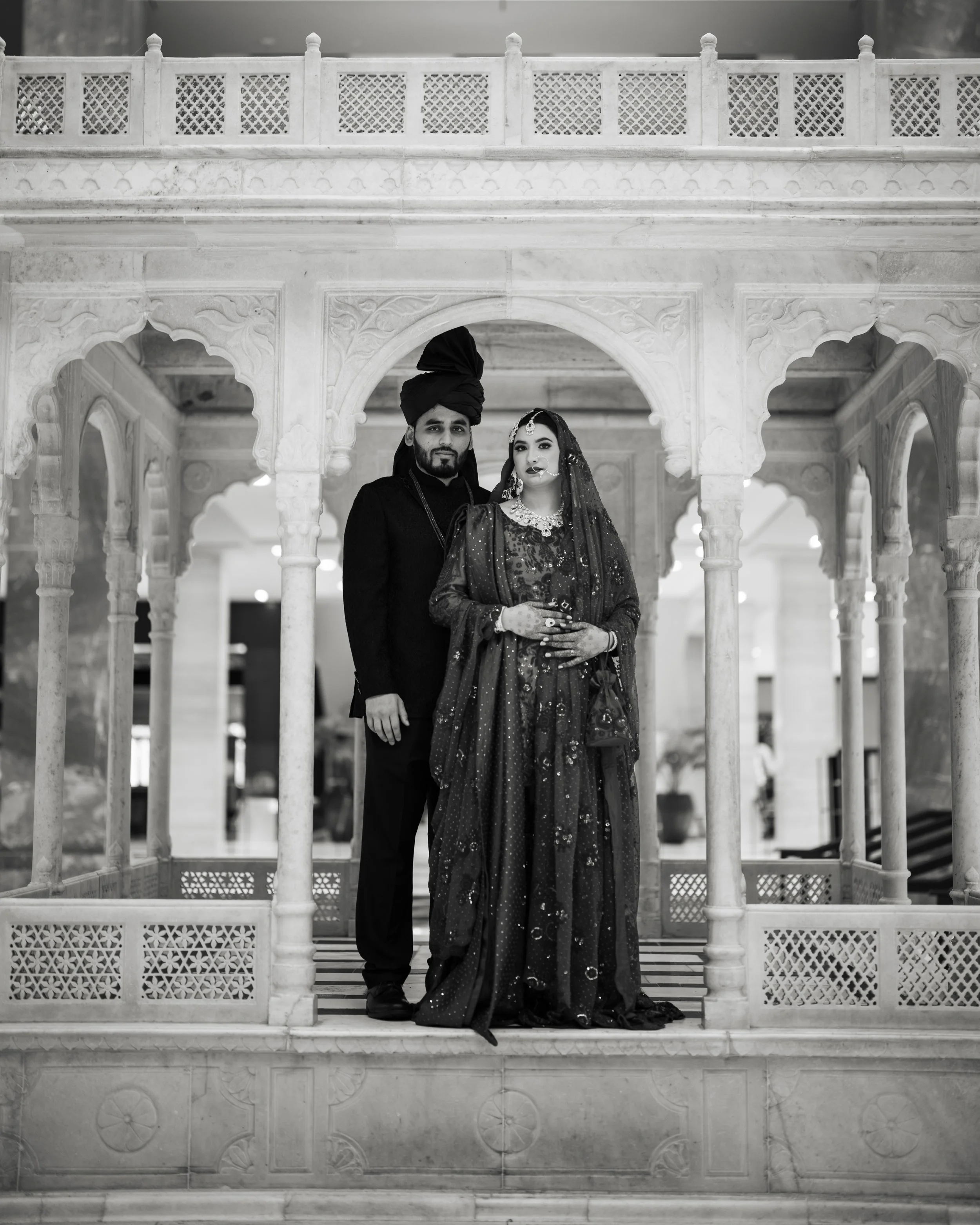 A couple dressed in traditional Indian wedding attire standing inside an ornate marble pavilion. The man is wearing a dark sherwani and turban, while the woman is dressed in a heavily embellished sari with jewelry and a veil. They are posing together