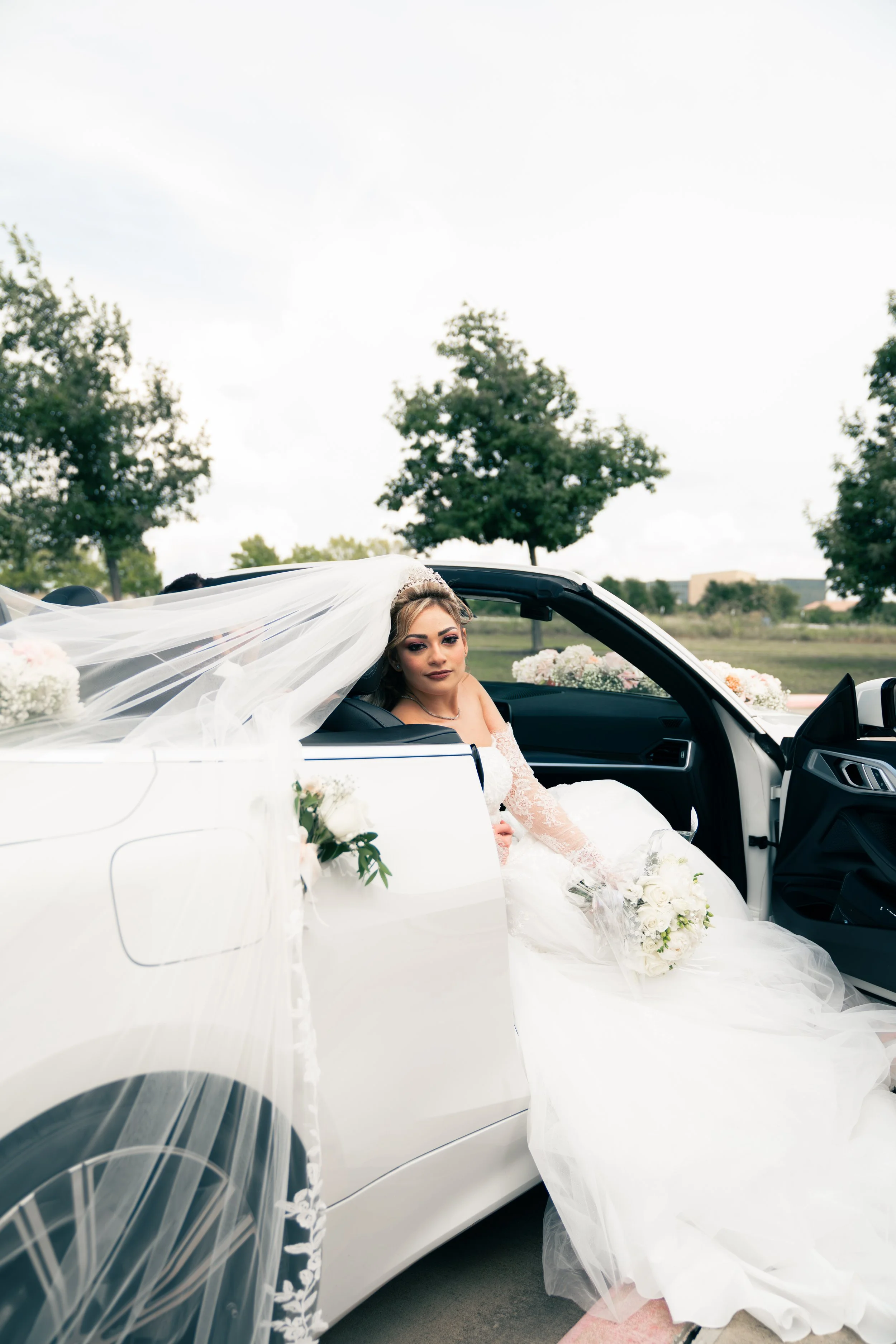 Bride in a wedding dress sitting in a white convertible car decorated with flowers, holding a bouquet, with a veil flowing behind her, outdoors with trees in the background.