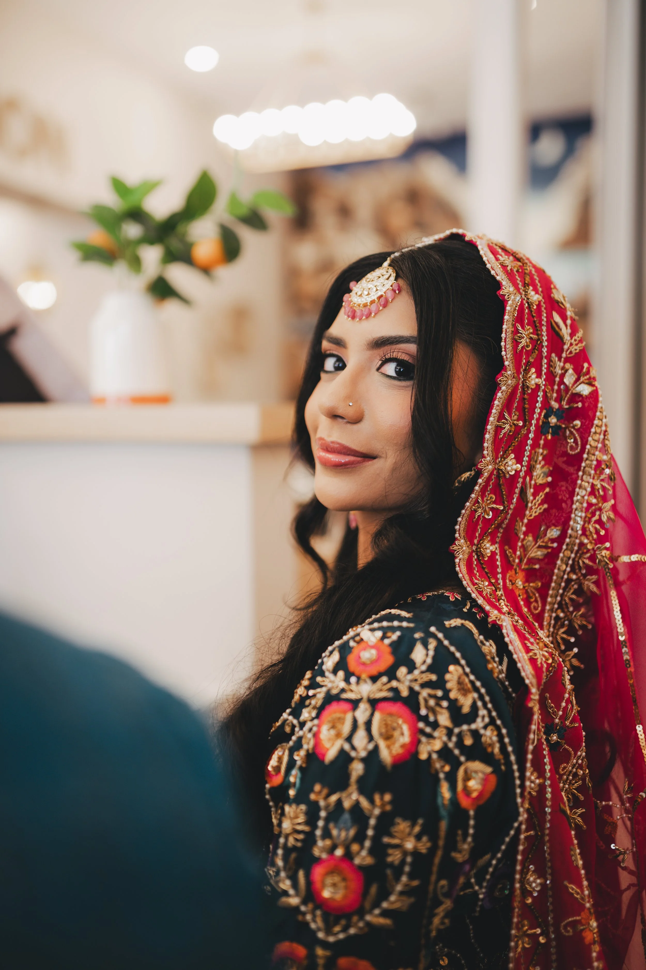 A woman in traditional Indian attire, wearing a red embroidered dupatta and jewelry, with dark hair and makeup, smiling softly in an indoor setting with bright lighting.