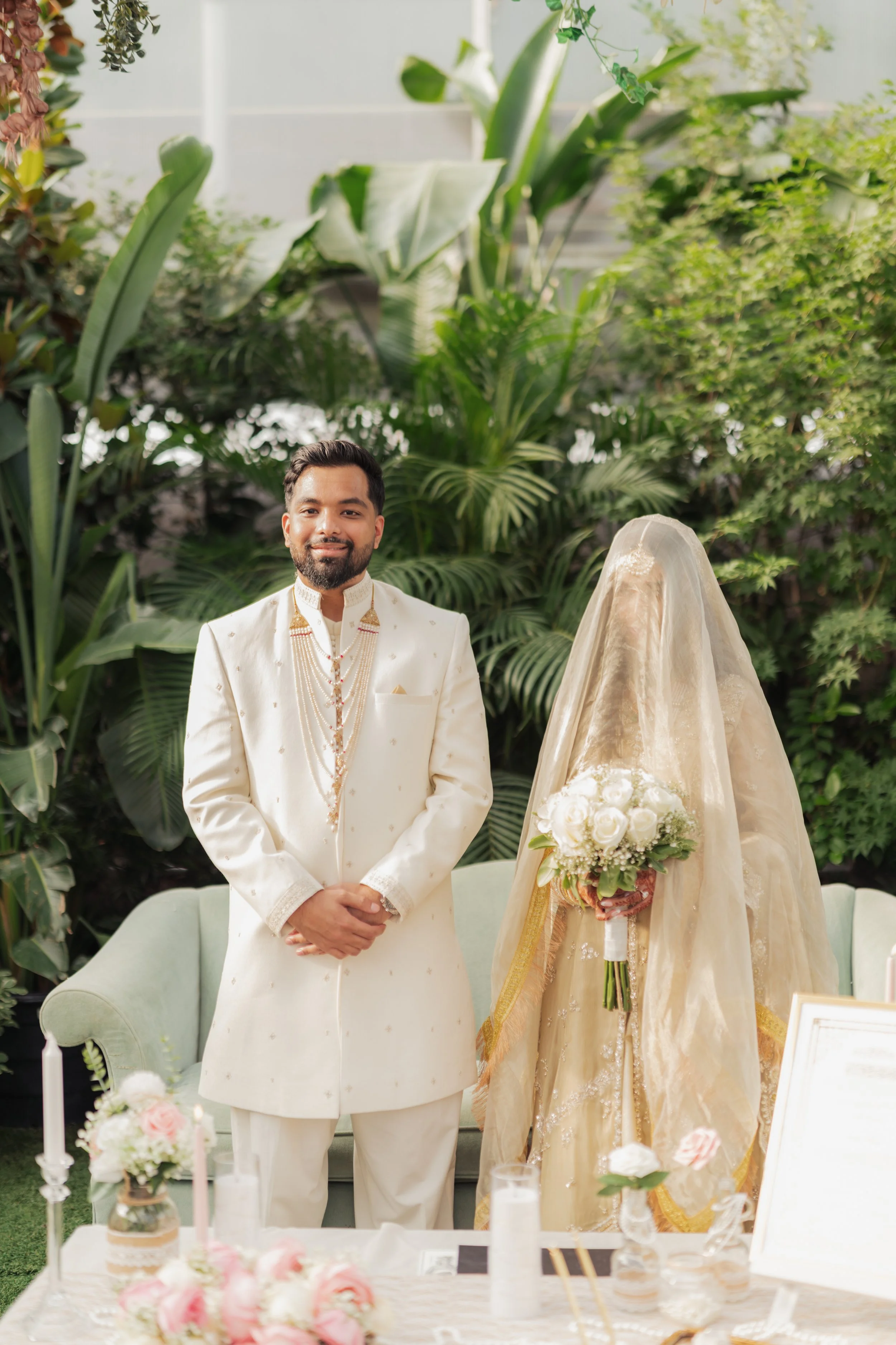 A man dressed in traditional South Asian wedding attire standing next to a bride in a gold wedding dress with a veil, holding a bouquet of white roses, in a lush garden setting with greenery and floral decorations.