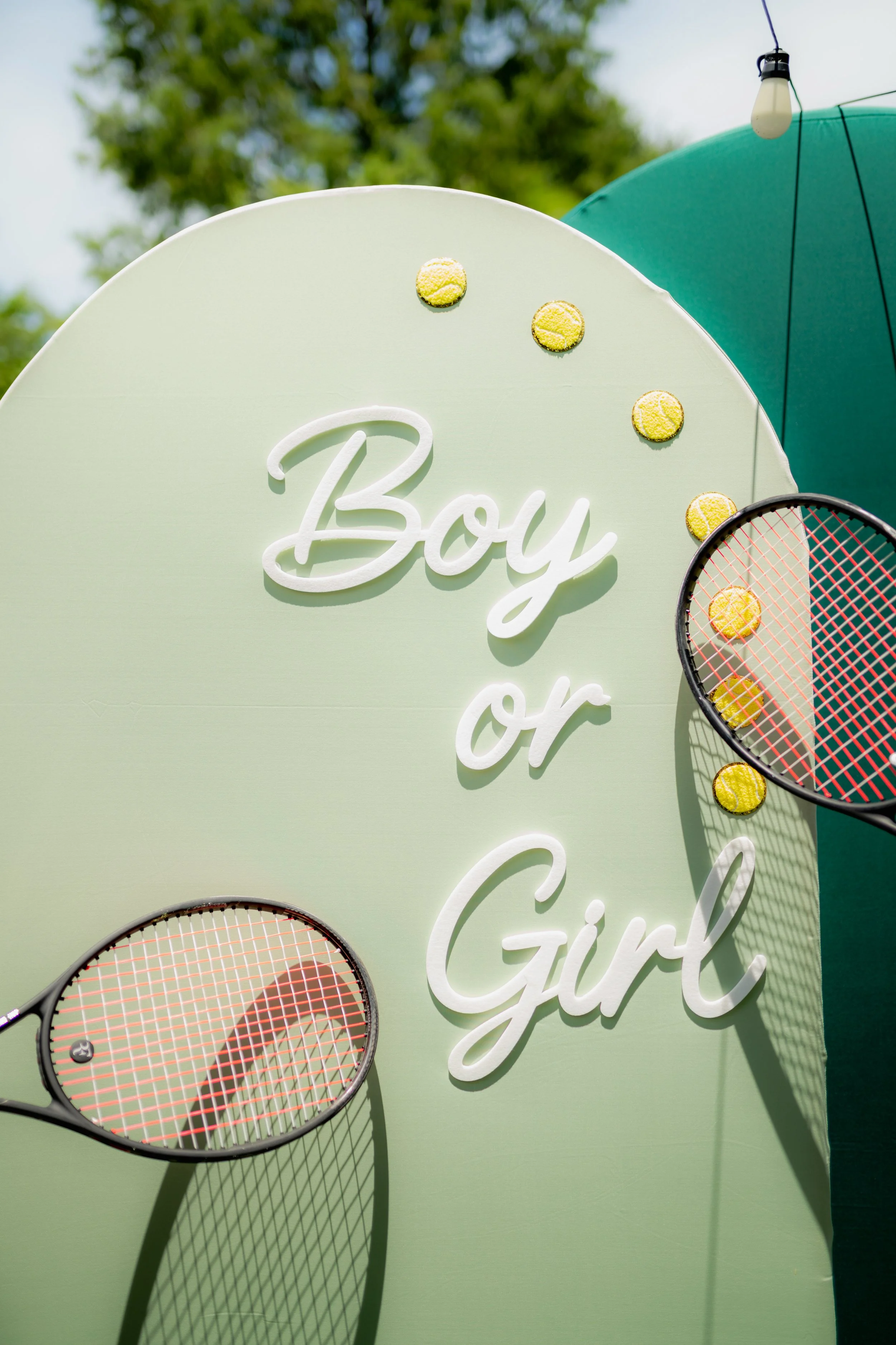 Decorative sign with tennis rackets, tennis balls, and the phrase 'Boy or Girl' against a light green background, with a teal umbrella and trees in the background.