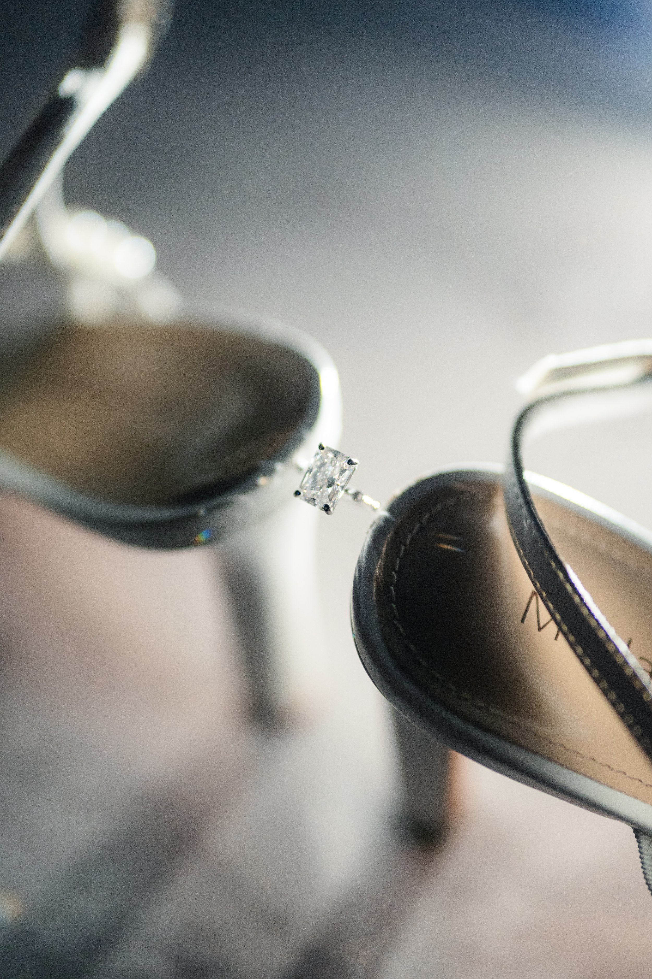 A diamond engagement ring between two black high-heeled shoes with silver accents, placed on a surface with soft lighting.