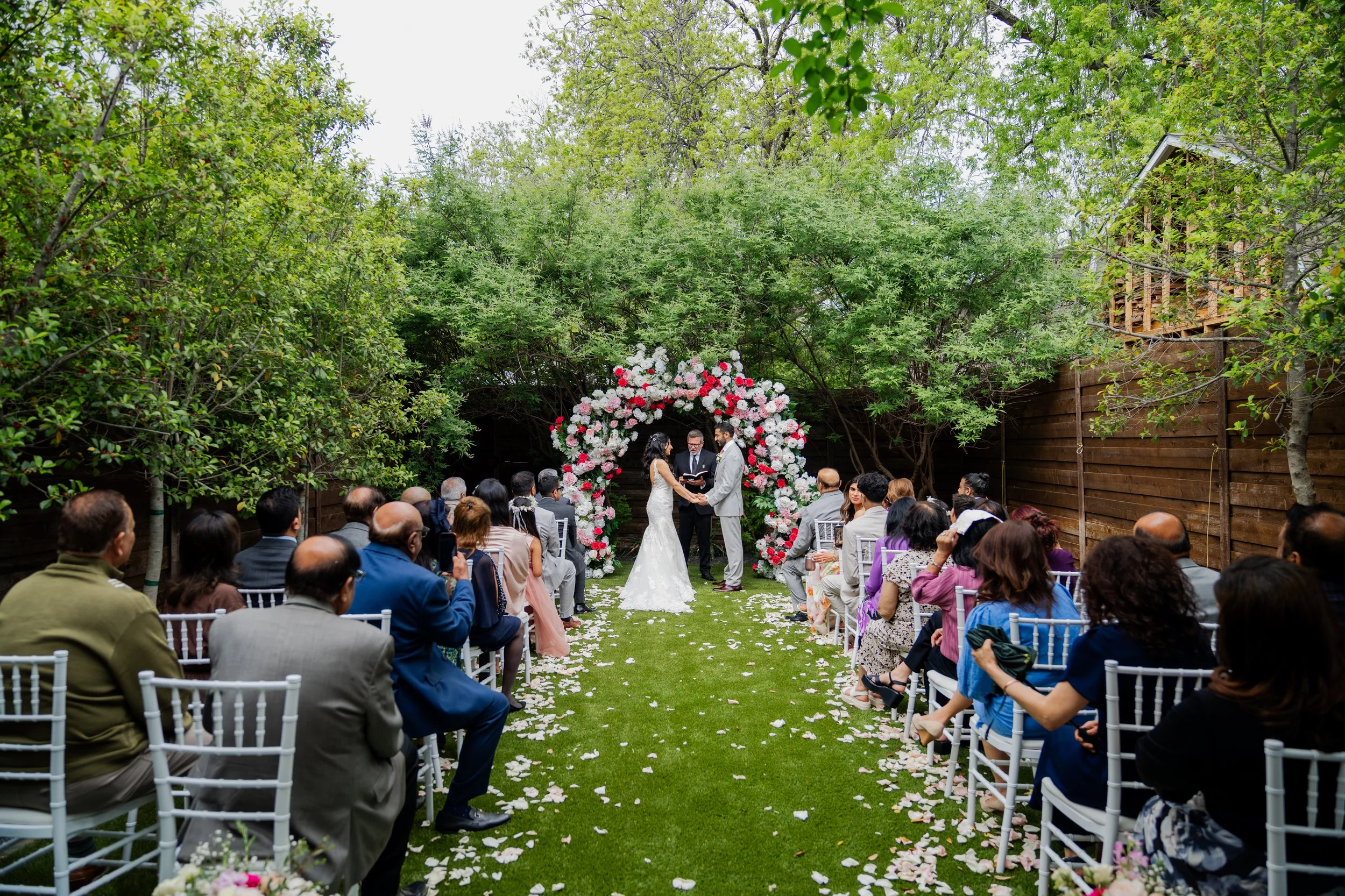 Bride and groom exchange vows under a floral arch at an outdoor wedding ceremony with seated guests, surrounded by trees and greenery.