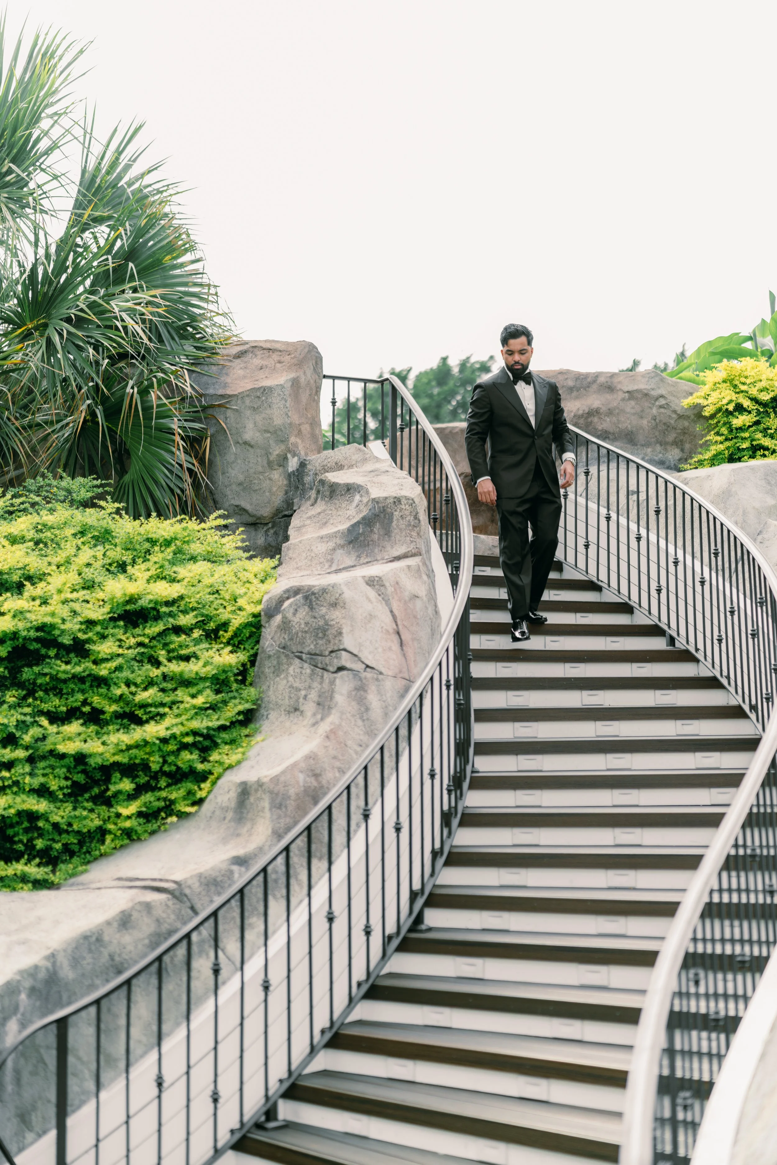 A man in a black tuxedo walking down an outdoor staircase with black railings, surrounded by large rocks and green plants.