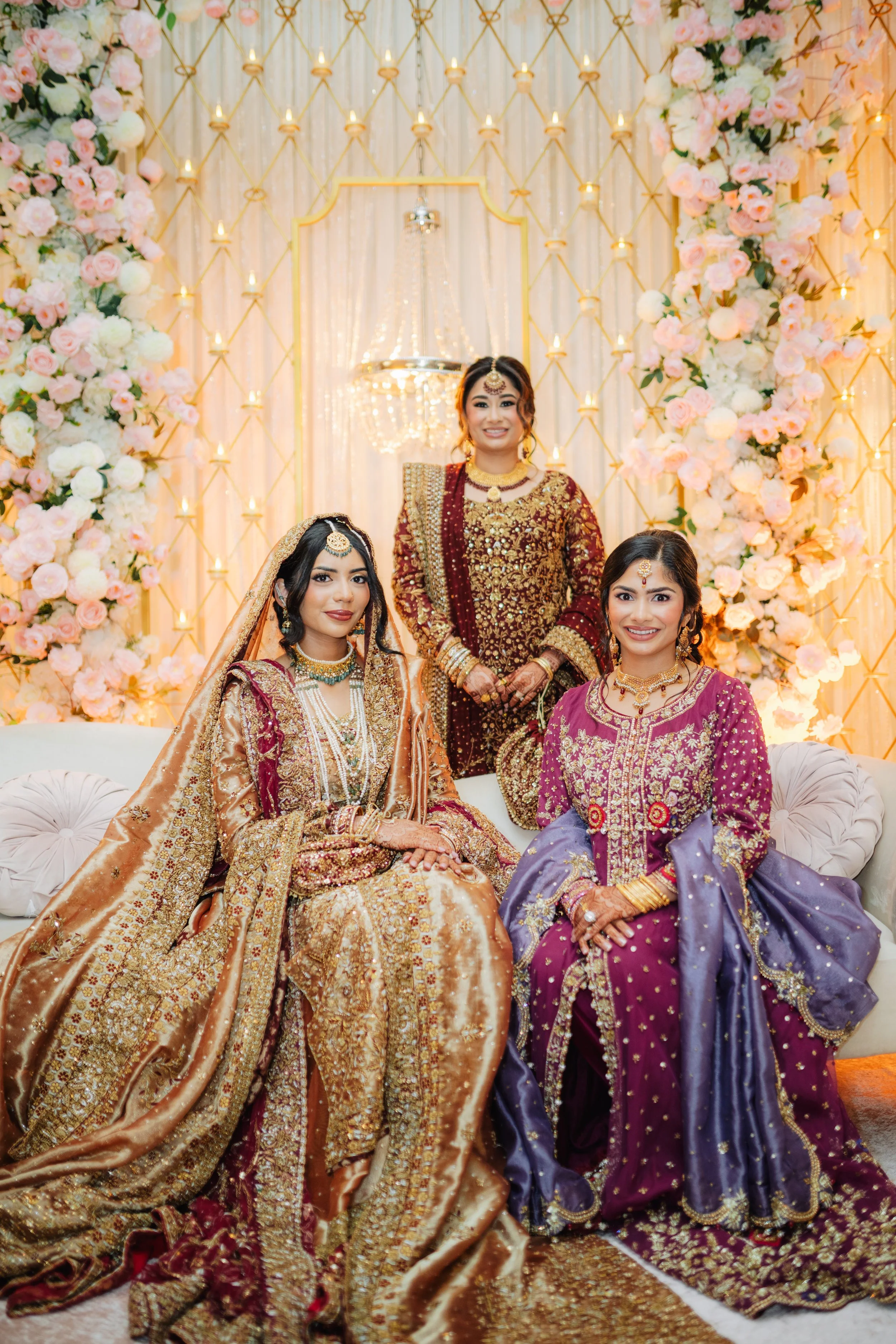 Three women dressed in traditional South Asian wedding attire sitting and standing in front of a floral and candle backdrop.