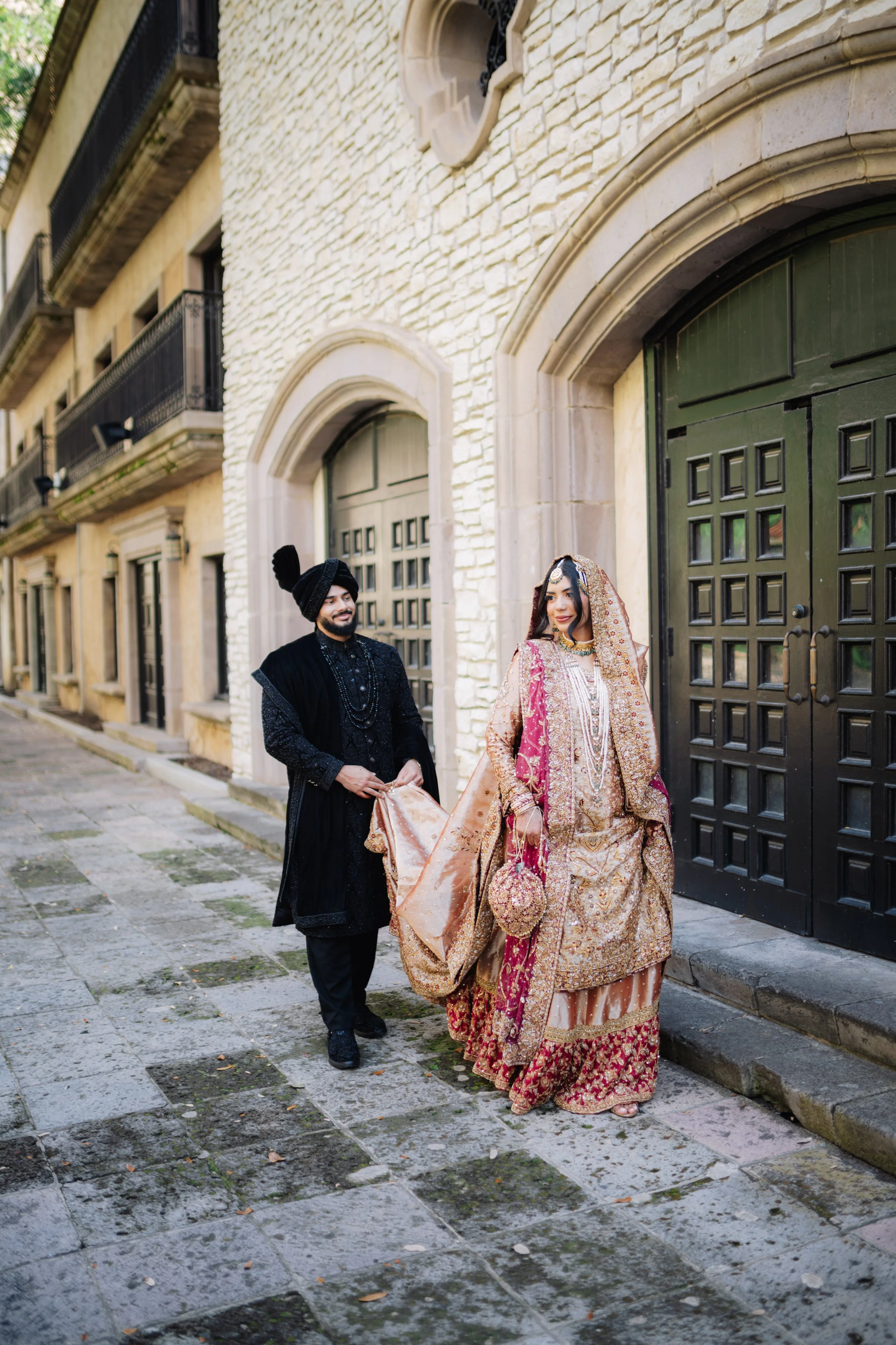 A South Asian bride and groom in traditional wedding attire walking past a stone building with black doors and balconies.