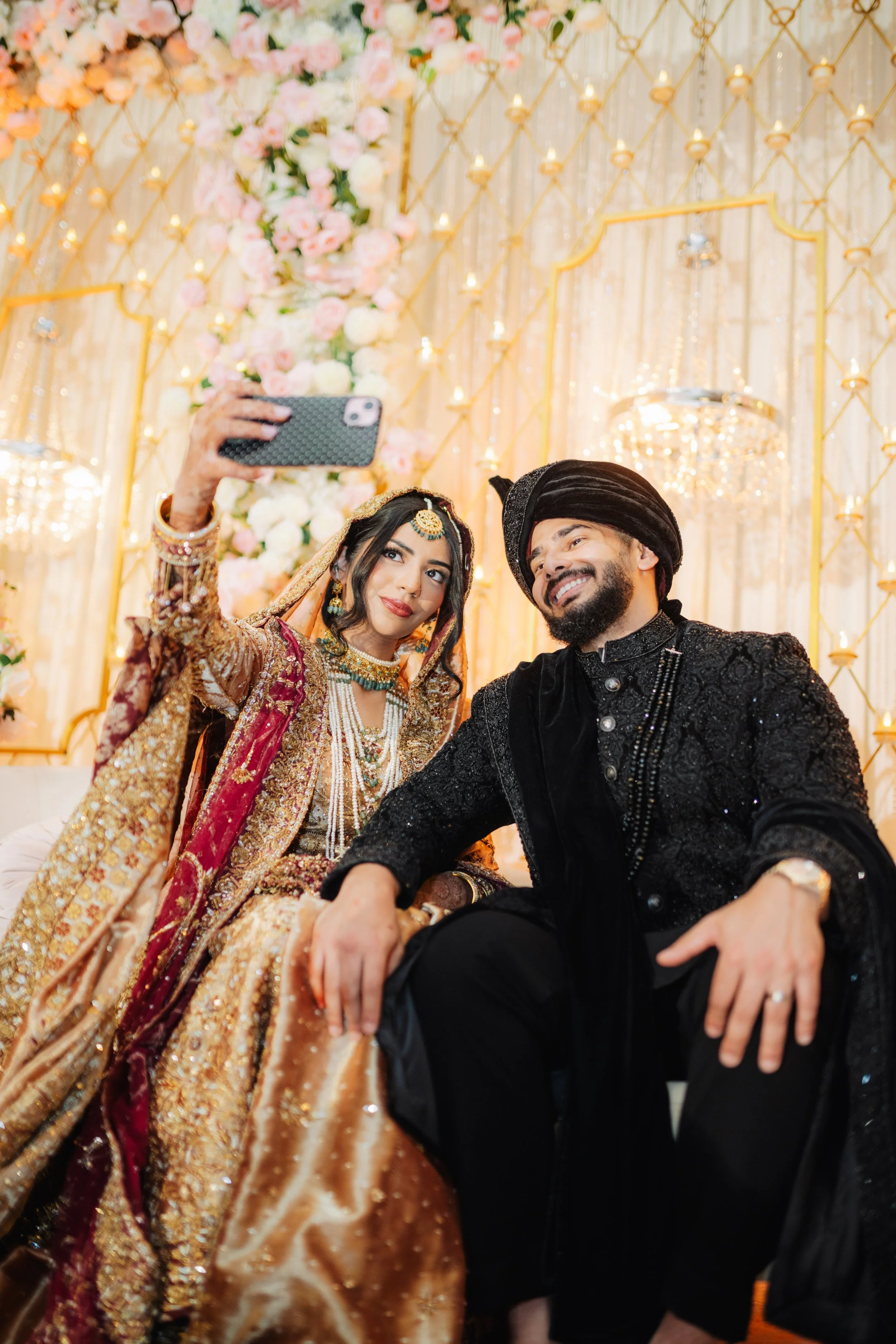 A bride and groom in traditional South Asian wedding attire taking a selfie together at their wedding celebration, with a floral and chandelier decorated background.