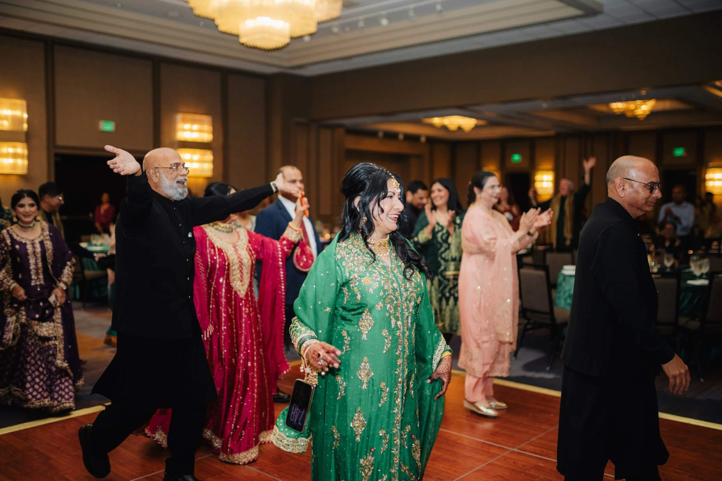 People dancing and celebrating at a formal event, with women in colorful traditional dresses and men in black and dark suits, in a ballroom with chandeliers.