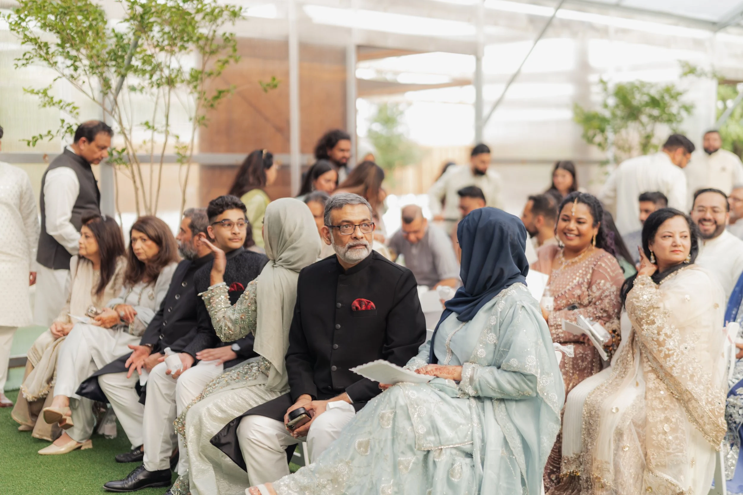 Group of people attending a ceremony, many dressed in formal or traditional South Asian clothing, seated in rows inside a bright, modern venue with glass walls and trees.
