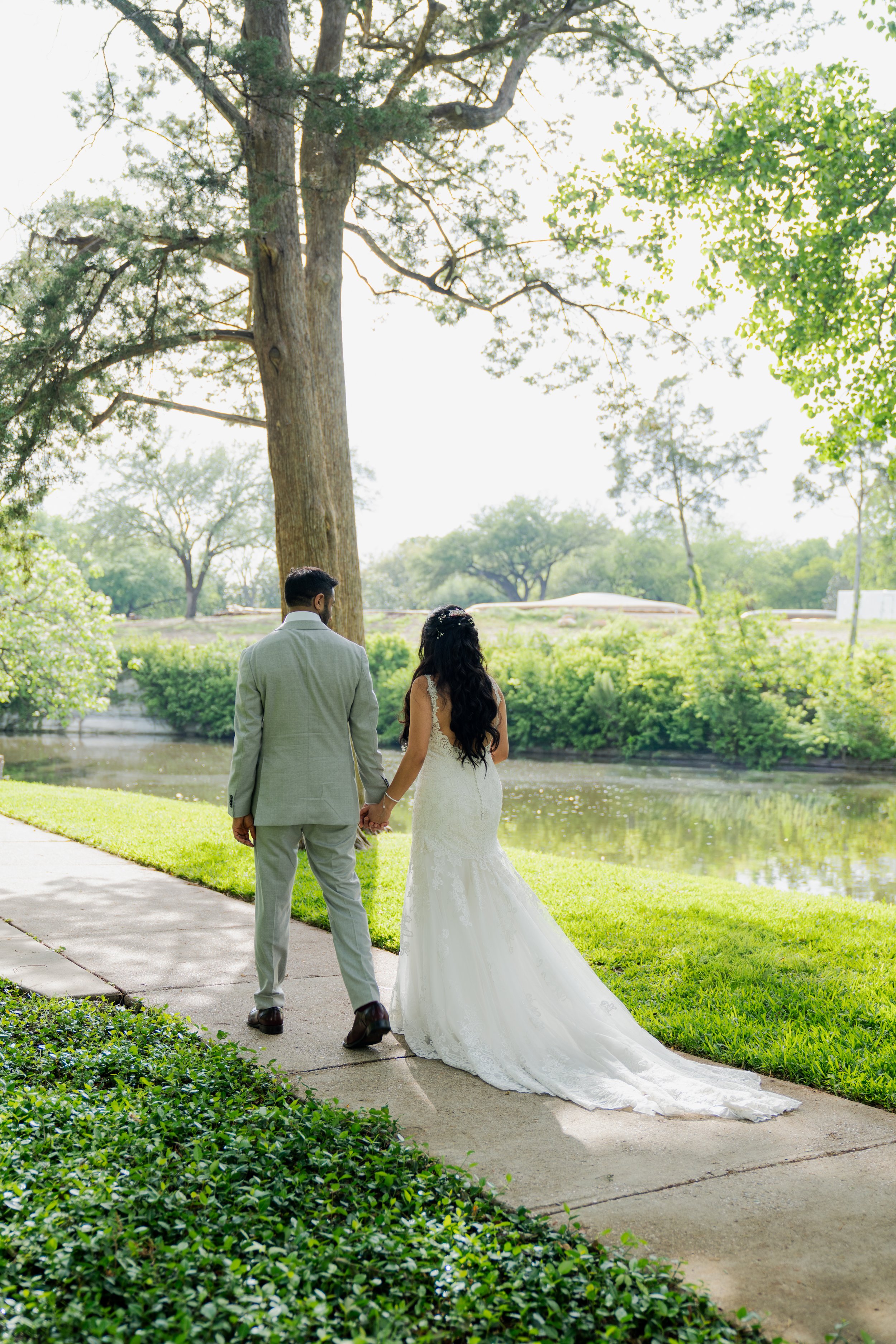 A bride and groom holding hands as they walk along a park path near a river, surrounded by green trees and shrubbery, with the bride wearing a white wedding gown and the groom in a light gray suit.