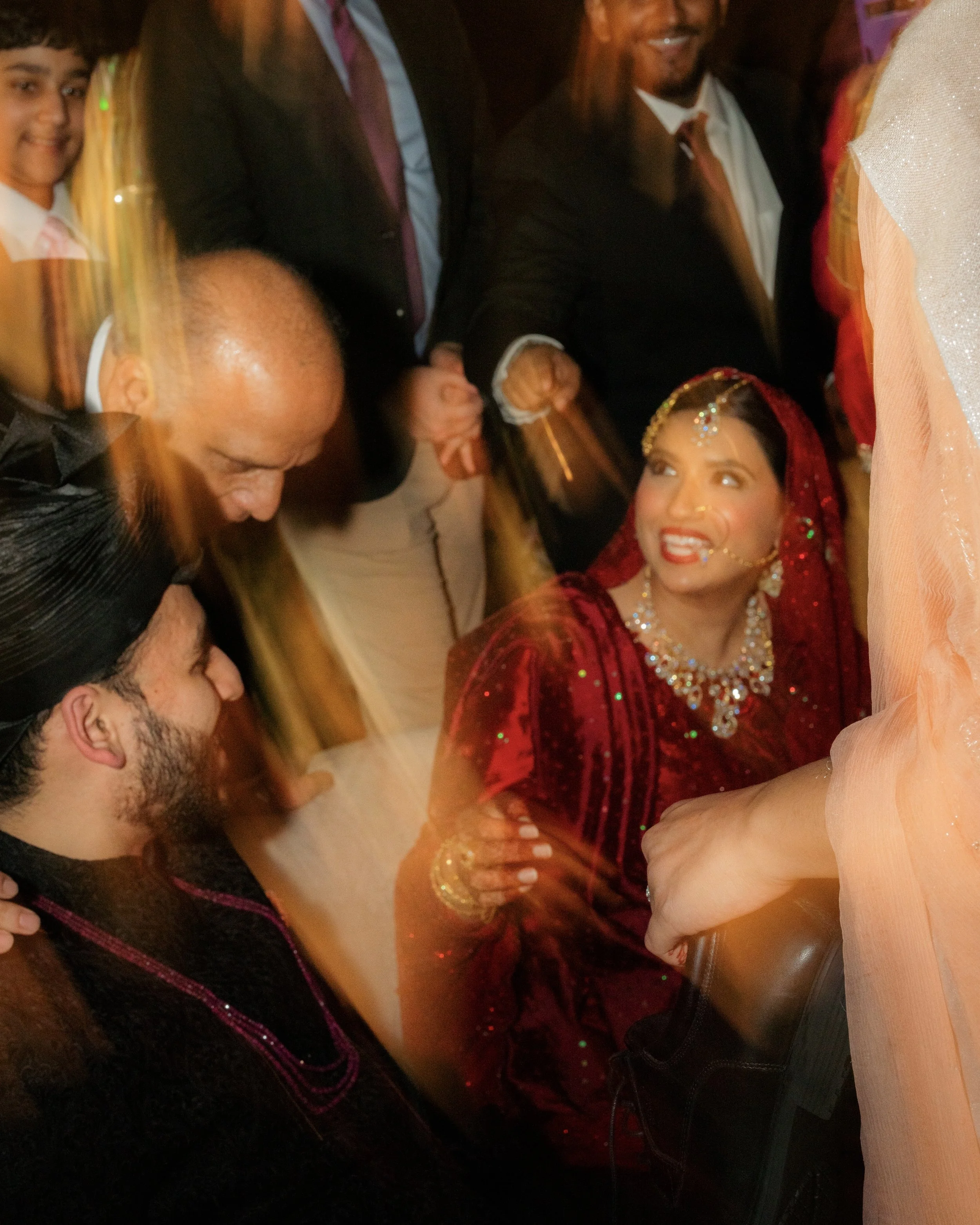 A woman dressed in a red traditional outfit with jewelry is sitting and interacting with men dressed in formal attire, surrounding her at a celebration or wedding.