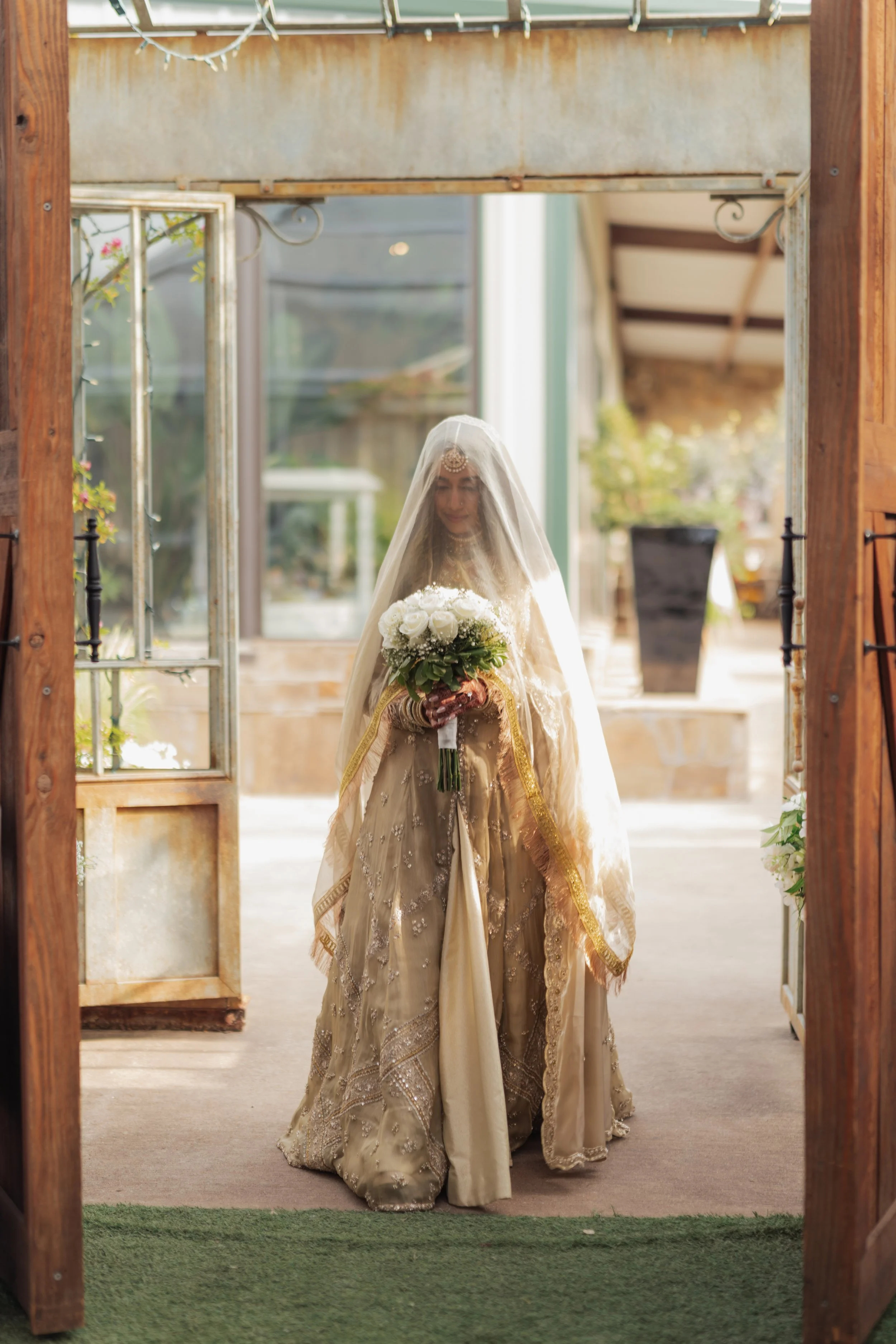 A woman dressed in traditional bridal attire holding a bouquet of white roses and wearing a veil, standing under an archway made of wood, with sunlight streaming through. The scene appears to be at a wedding or special ceremony.