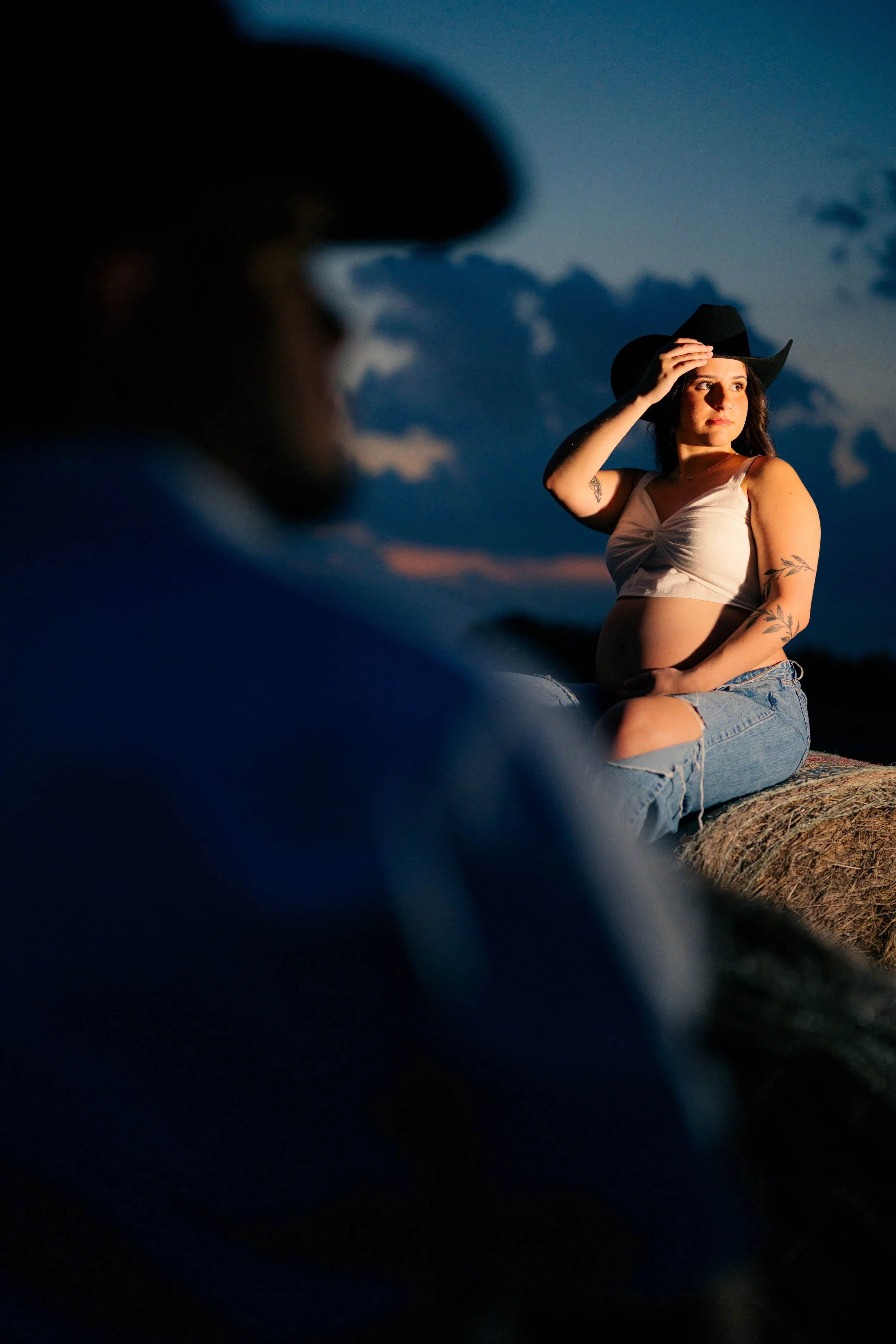 A young woman wearing a cowboy hat, a white crop top, and ripped jeans sitting on a hay bale outdoors at dusk, with a darkened silhouette of a man in the foreground and a cloudy sky in the background.