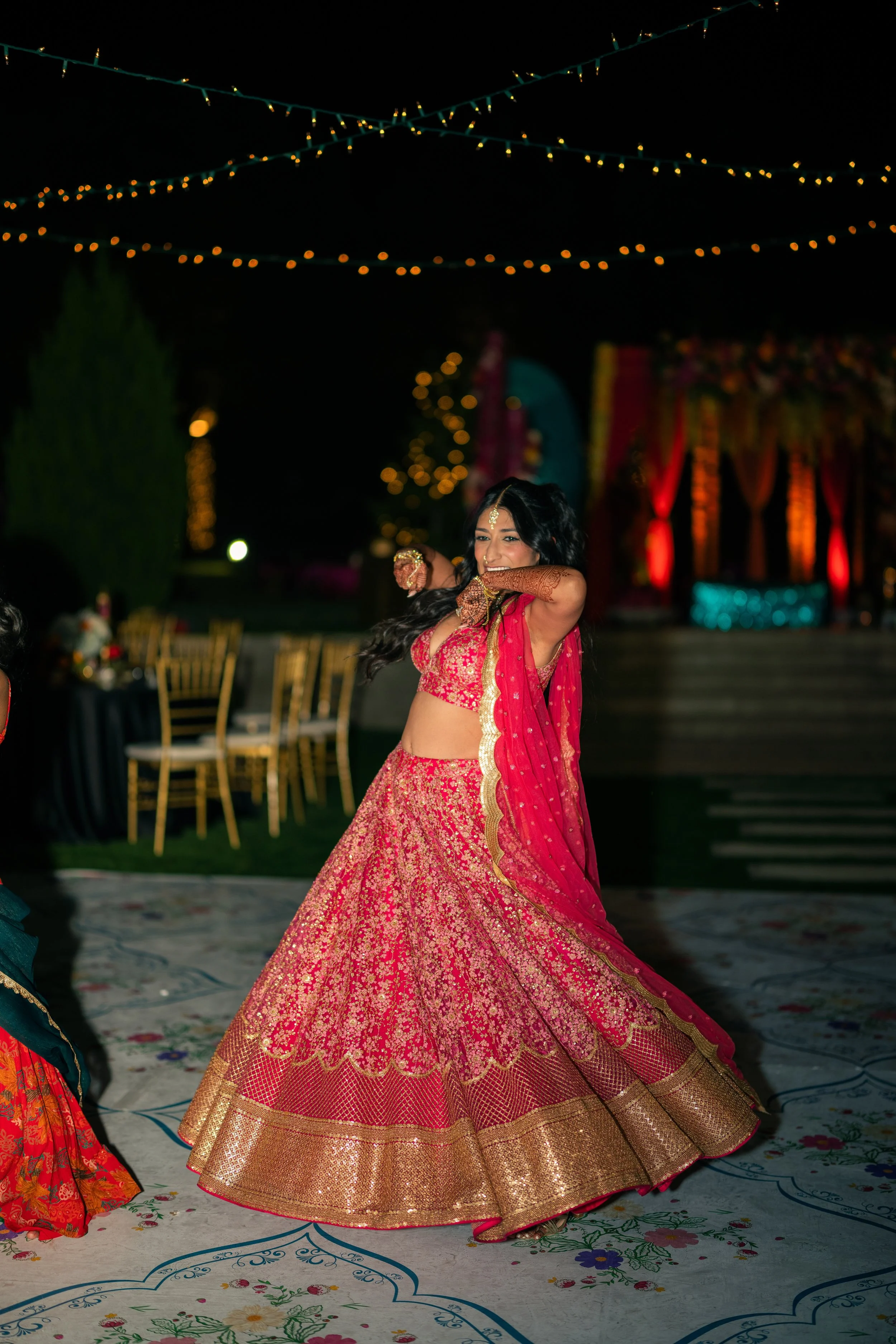 A woman in a pink and gold traditional Indian outfit dancing at a nighttime celebration outdoors, decorated with string lights and colorful drapes.