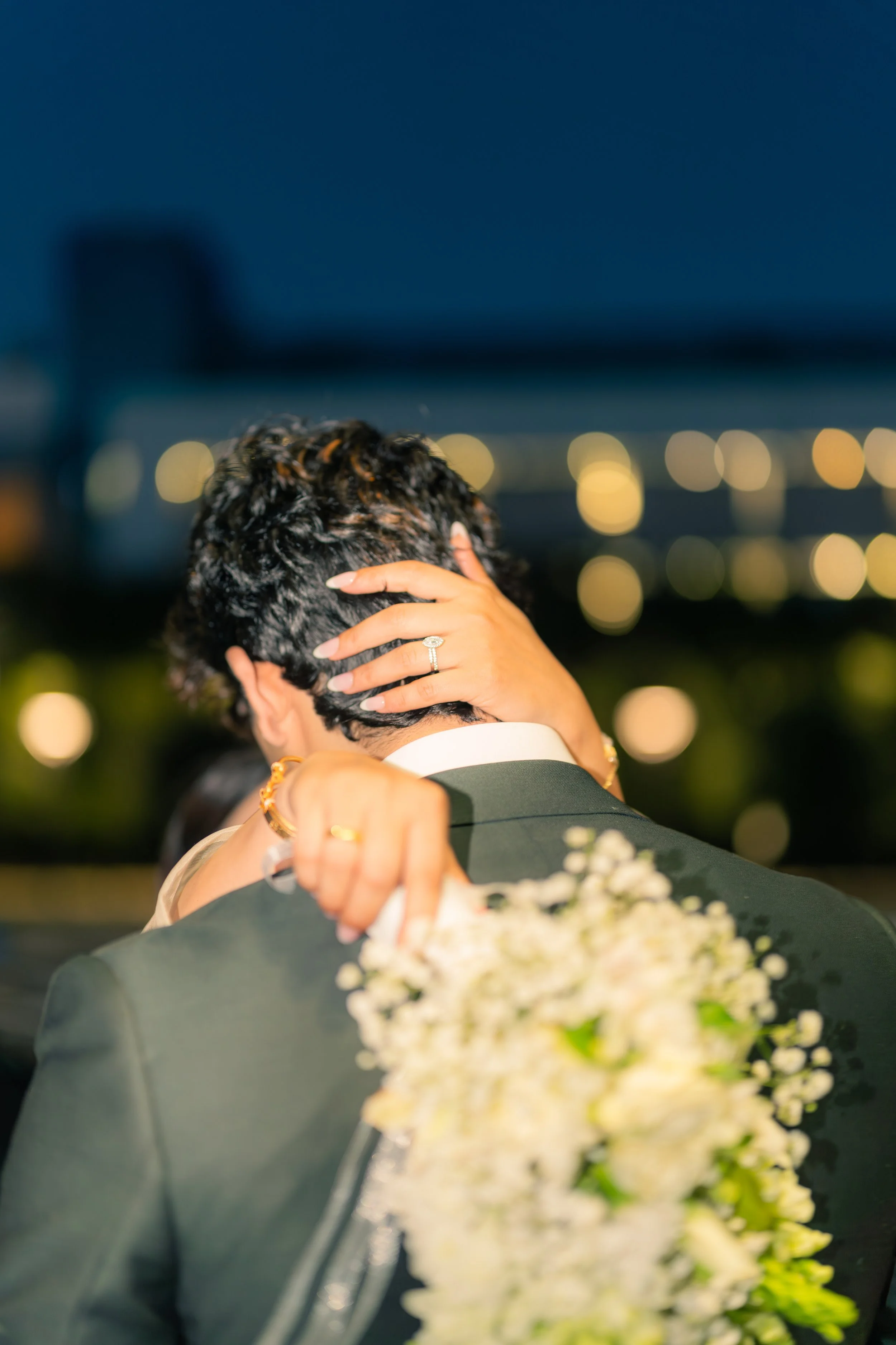 A couple sharing a romantic moments on their wedding day at night, with city lights in the background, embracing each other closely.