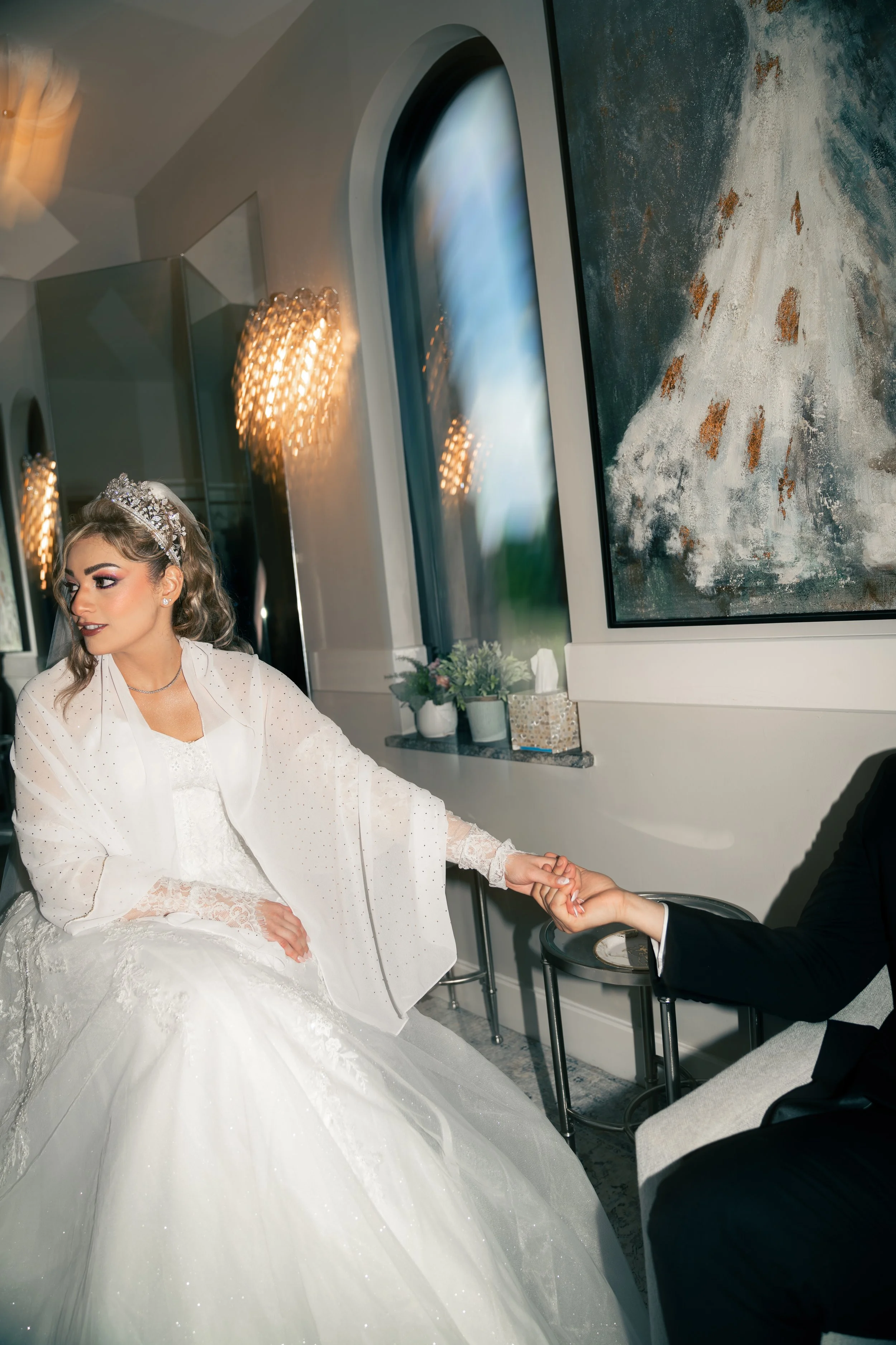 A bride in a white wedding gown and sparkling tiara sitting on a chair, holding hands with a person in a black suit.