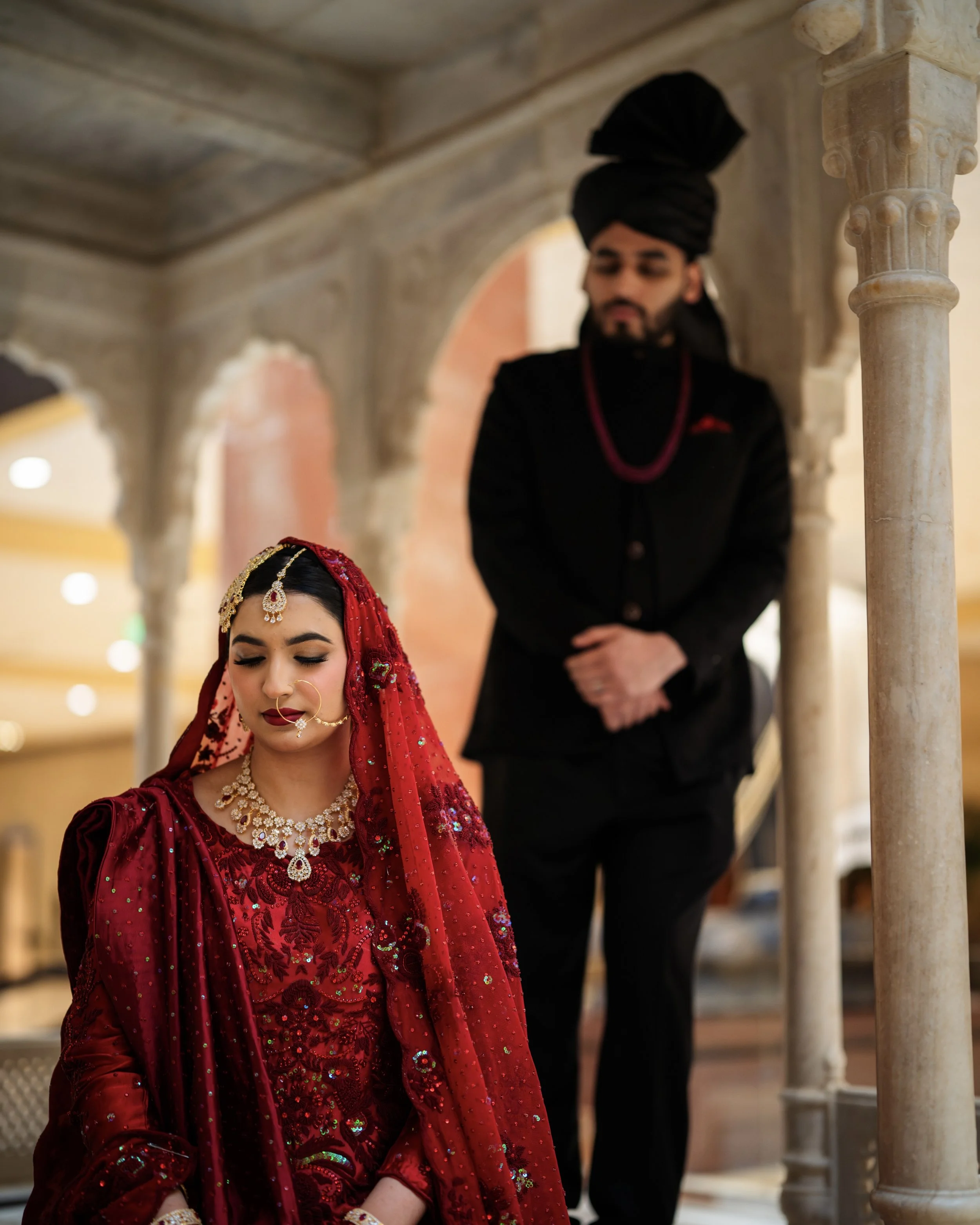 A bride in traditional red wedding attire and jewelry sits with her eyes closed, while a groom in black traditional attire stands behind her, looking down, in an ornately decorated indoor setting.