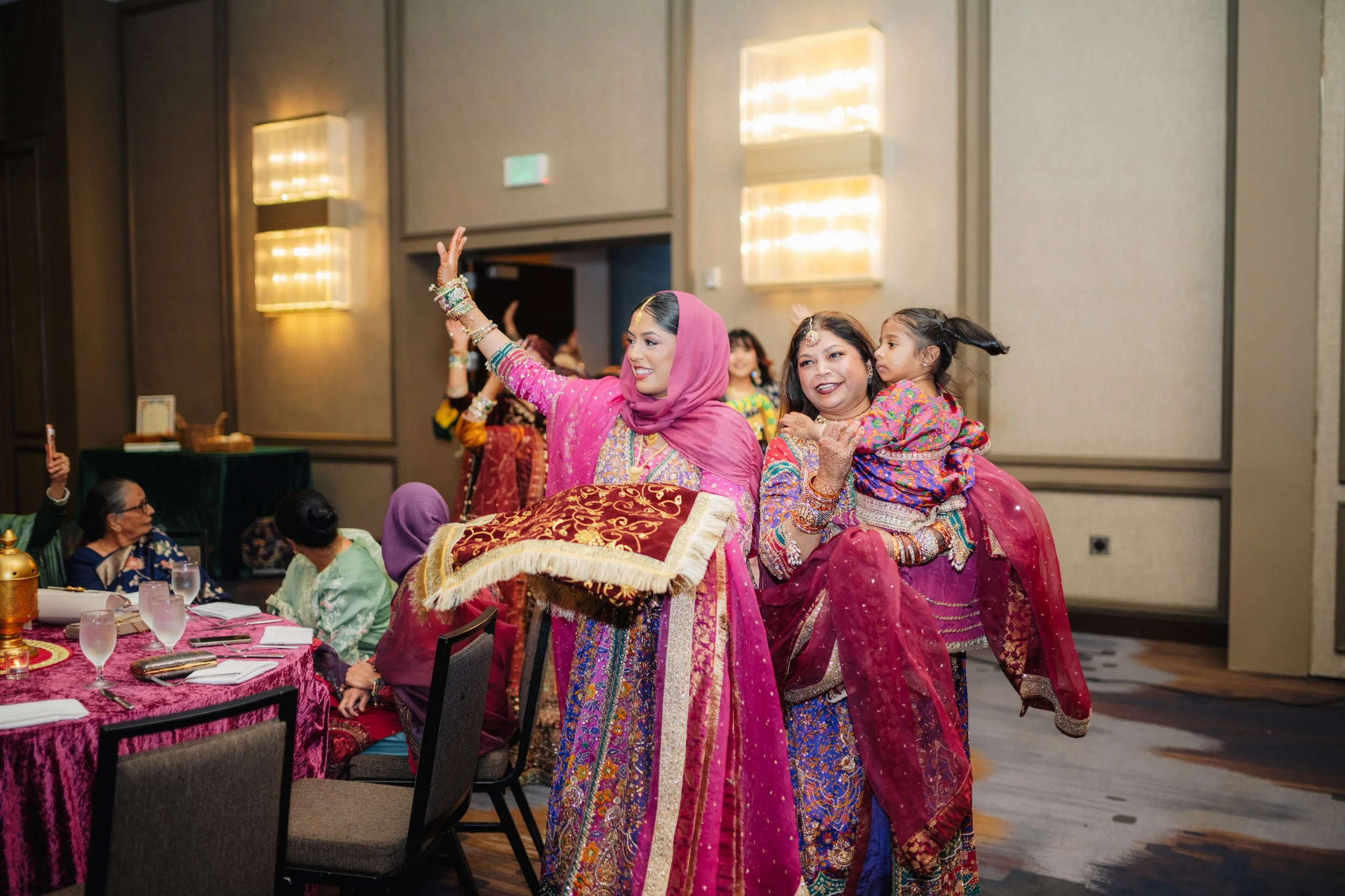 Women in colorful traditional South Asian attire dancing at a celebration event with seated guests watching.