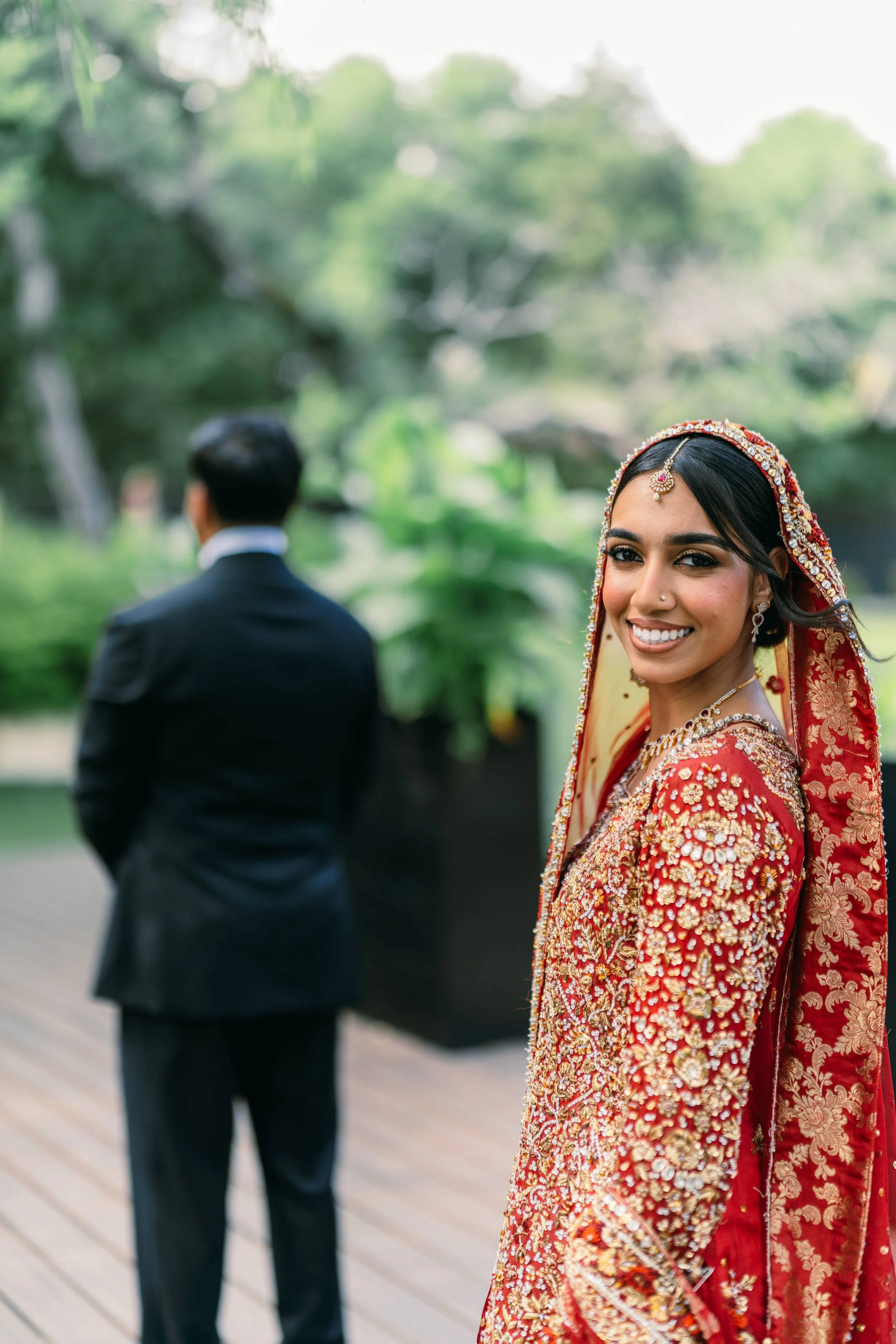 A smiling woman in traditional red and gold Indian wedding attire, standing outdoors with a woman in a black suit in the background.