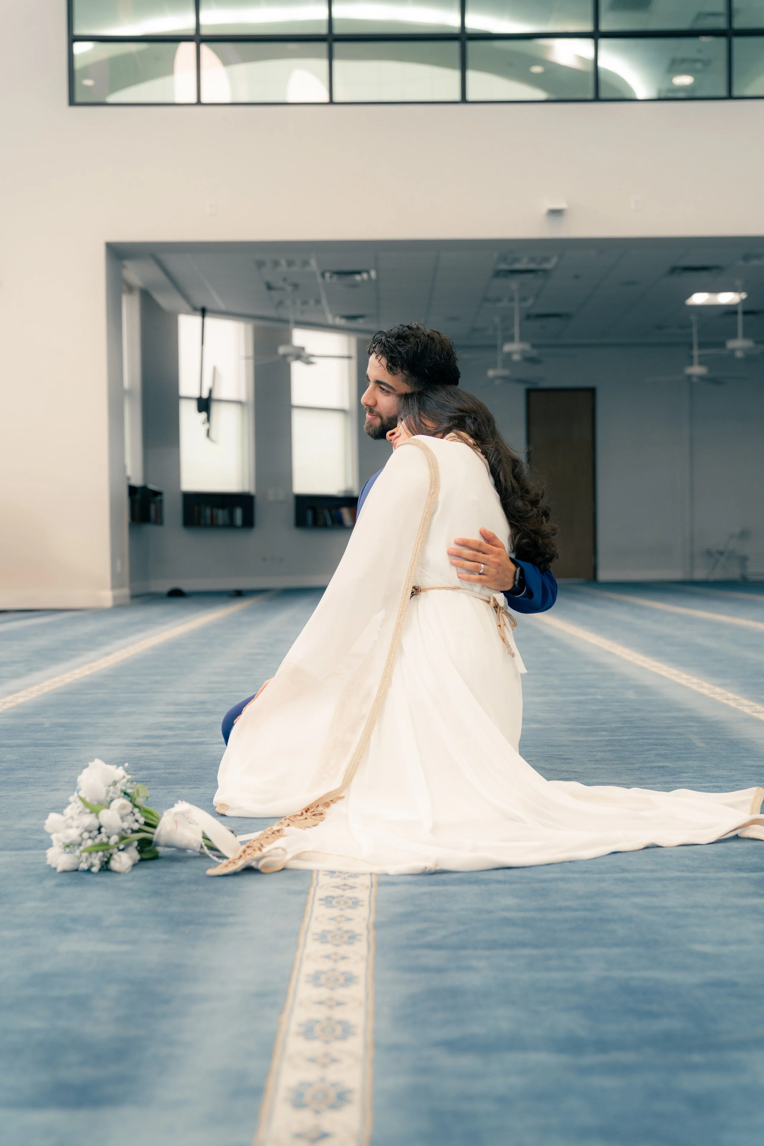 A couple in wedding attire embracing on a blue carpet inside a spacious, modern building with large windows.