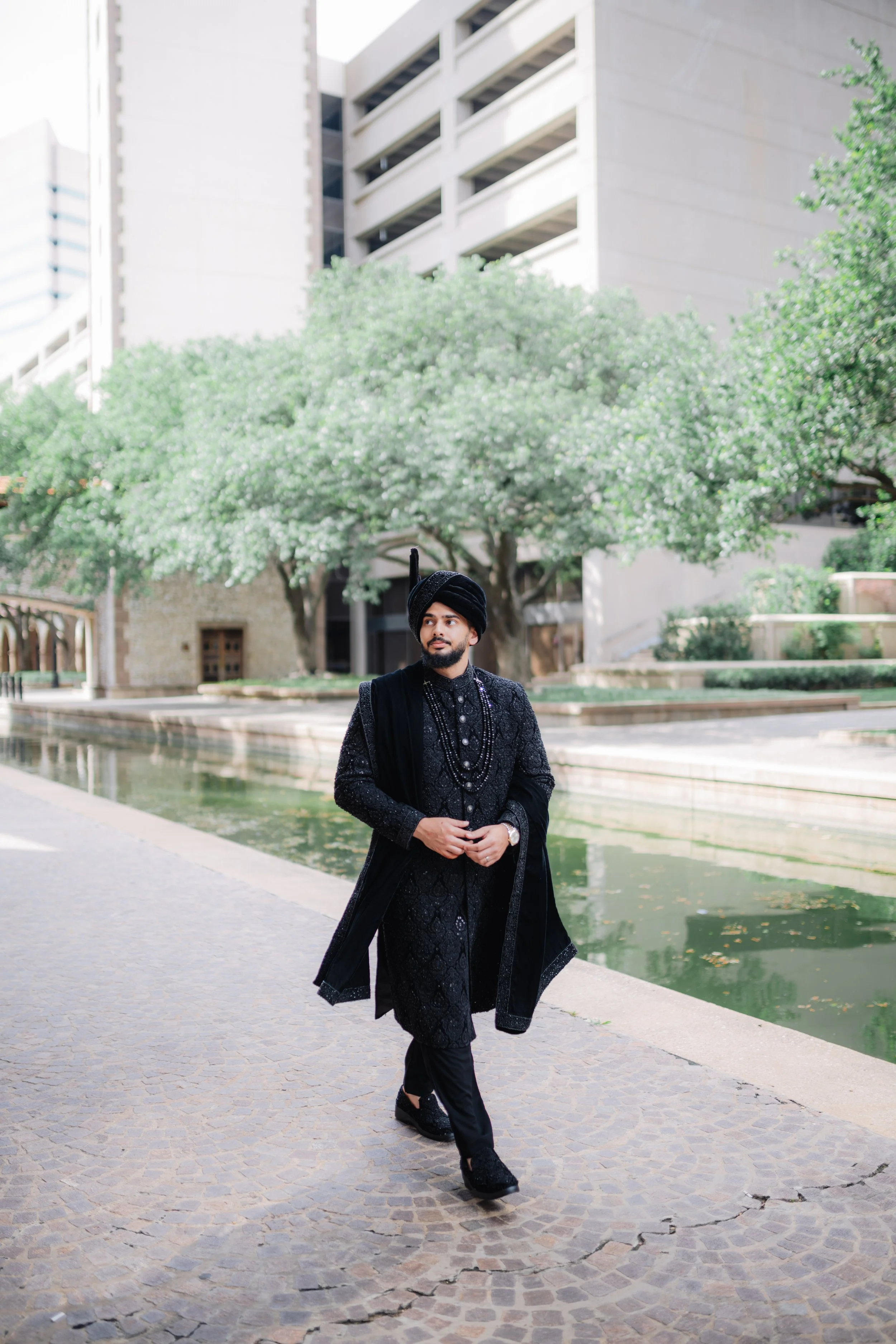 A man dressed in a black traditional outfit with a head covering and layered necklaces walking on a cobblestone path next to a narrow water canal, with modern buildings and green trees in the background.