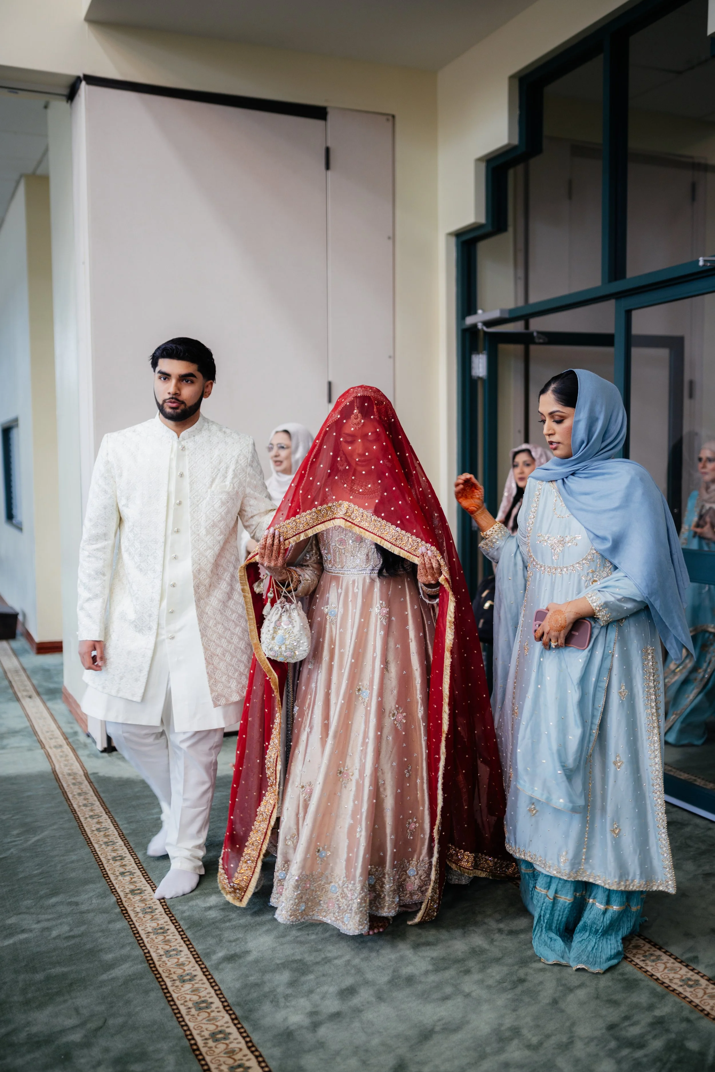 A group of South Asian people at a traditional wedding ceremony. The groom is wearing a cream-colored sherwani with white pants. The bride is dressed in a peach and gold lehenga with a red dupatta covering her head, holding a small bag. Two women, on