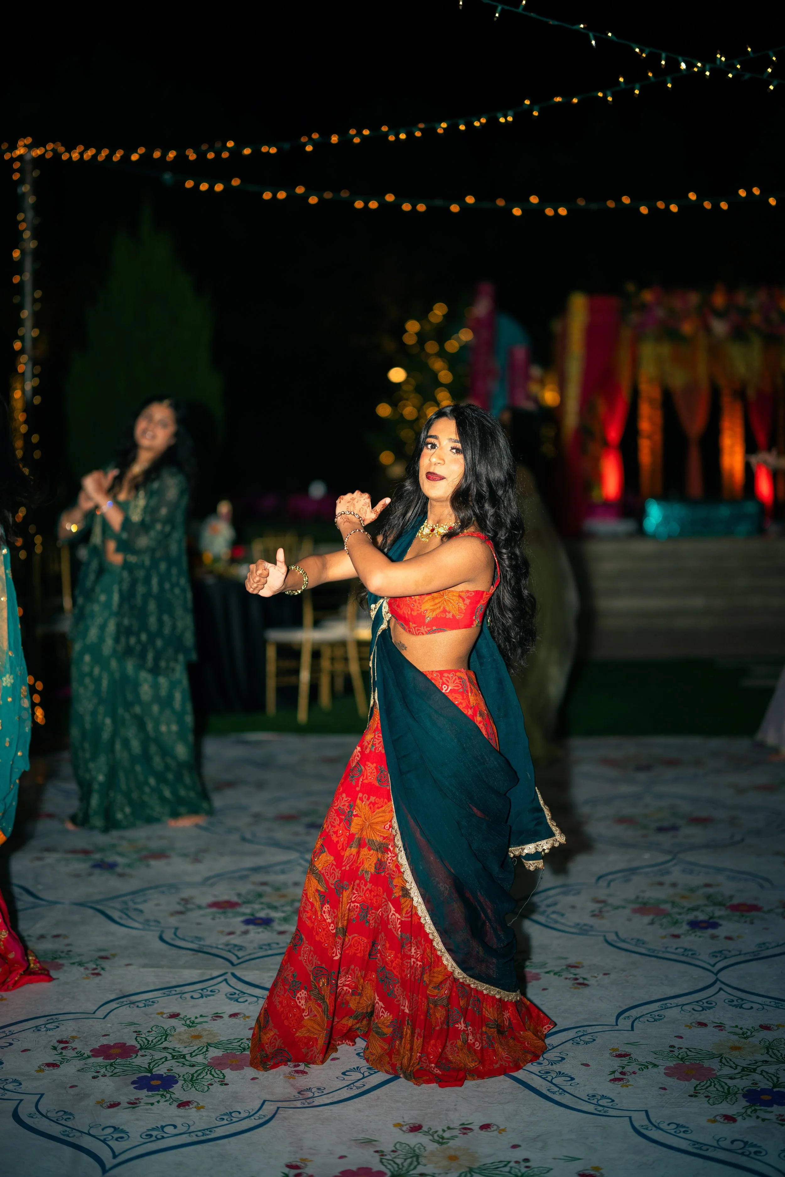 A woman in a red and blue traditional Indian outfit dancing at a nighttime celebration with string lights overhead and a decorated stage in the background.