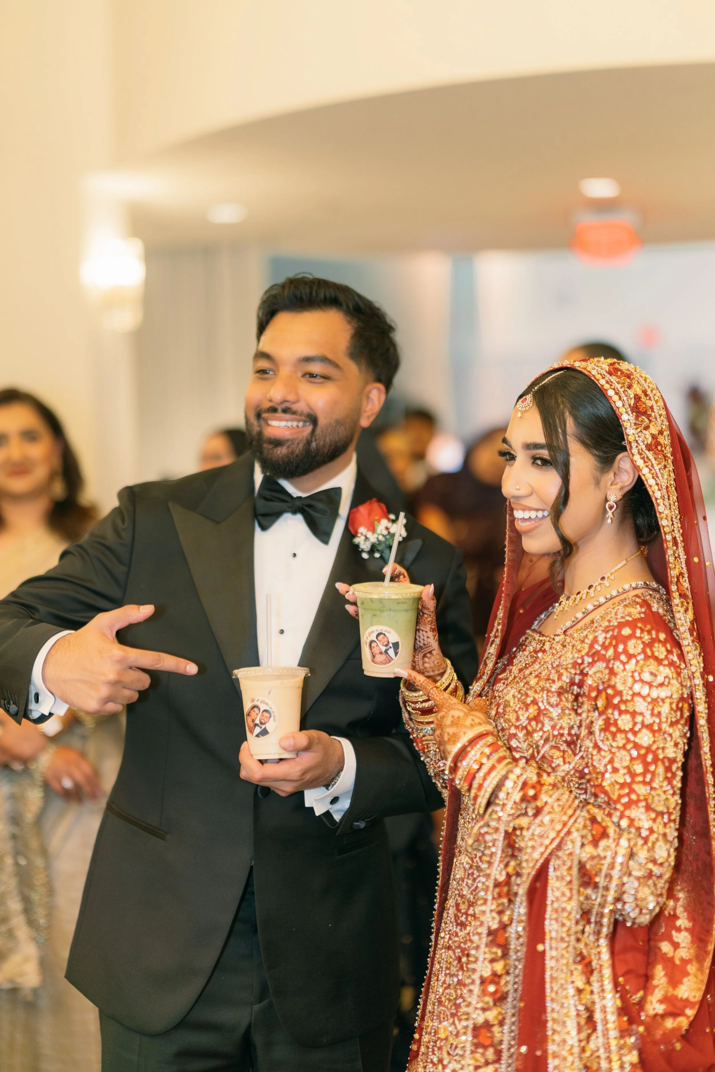 A man in a tuxedo and a woman in traditional Indian attire holding drinks at a social event.