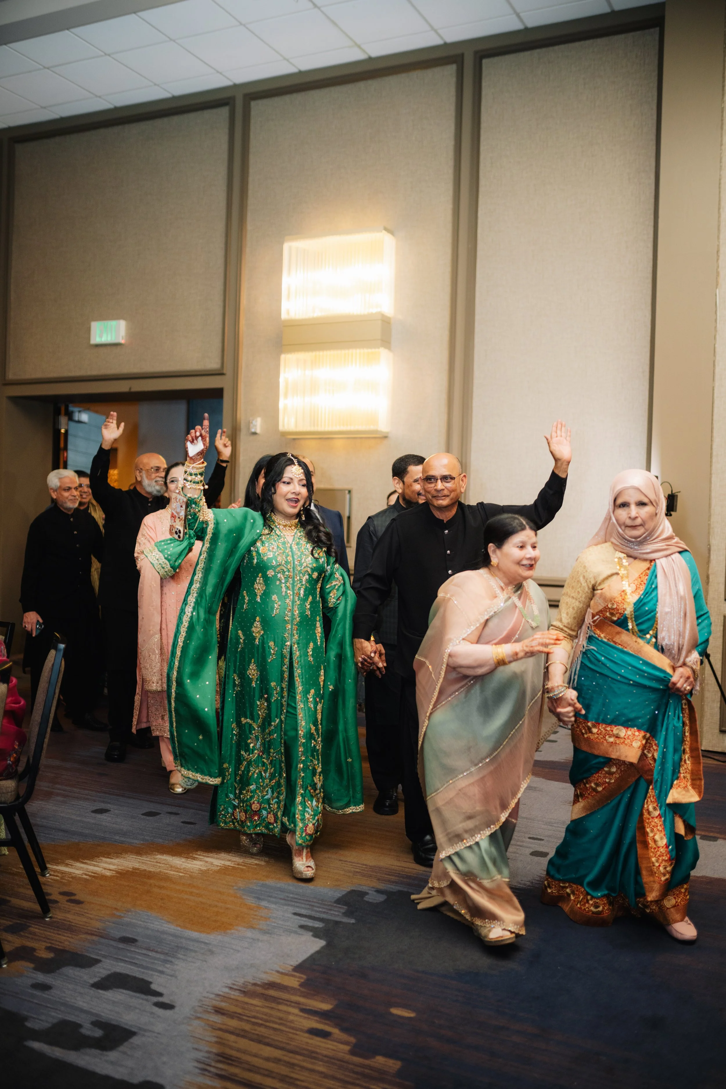 A group of people dressed in traditional South Asian clothing, walking and holding hands at an indoor event. The room has beige walls and a patterned carpet.