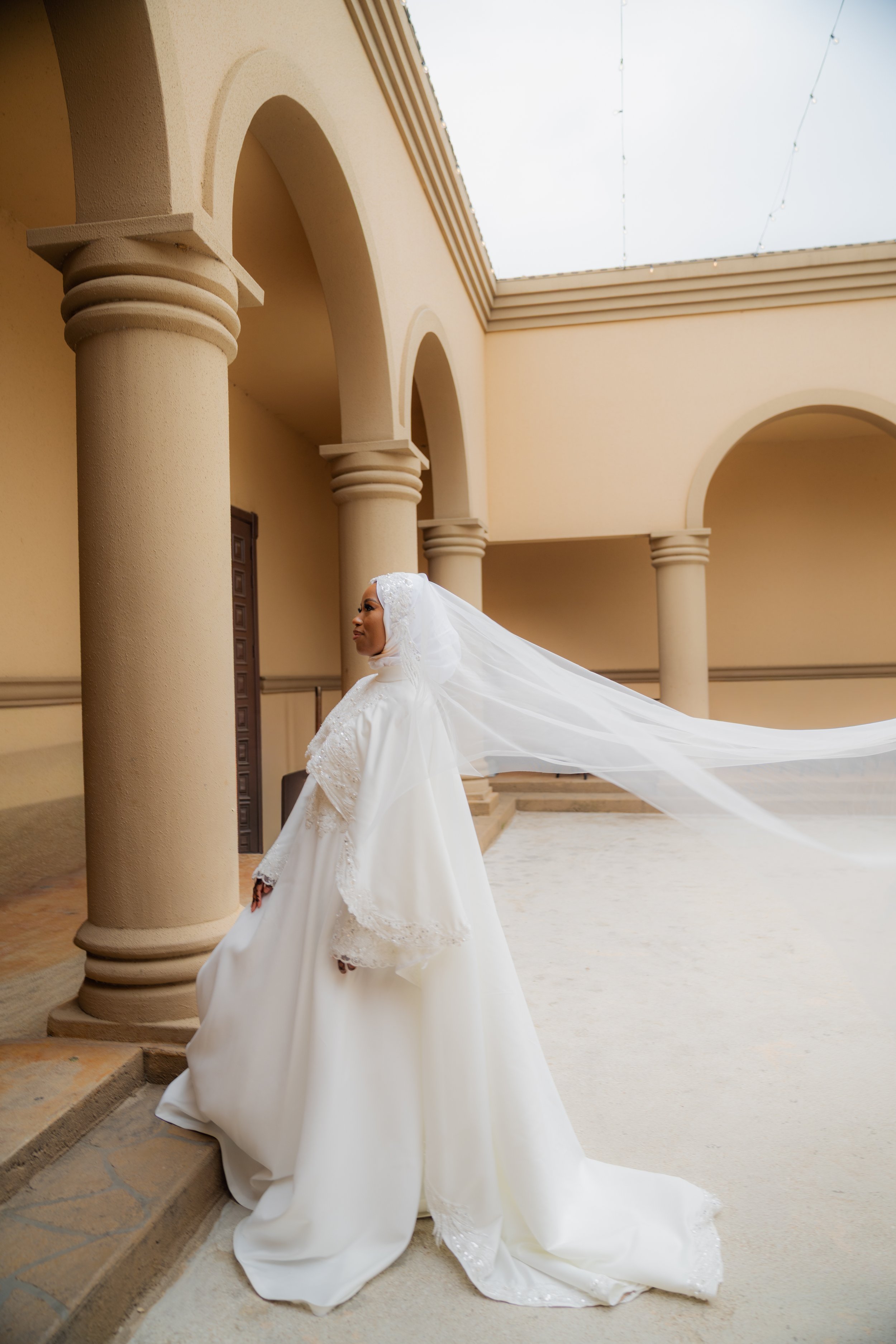 A woman in a white wedding dress with a veil flowing behind her stands on steps outside of a building with arched columns and beige walls.