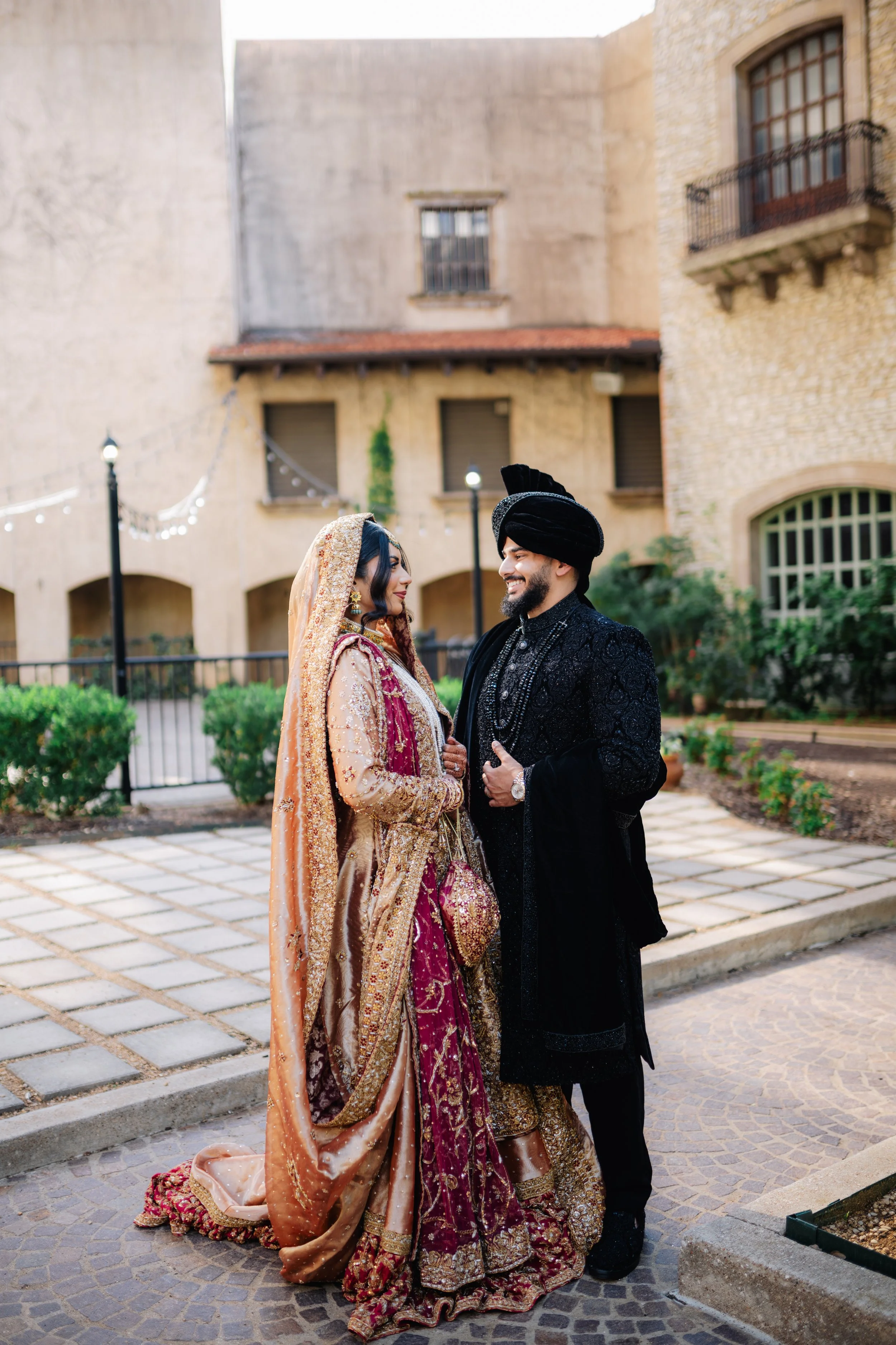 A bride and groom in traditional South Asian wedding attire standing outdoors in front of a building with brick and concrete walls. The bride is wearing a gold and maroon heavily embroidered dress with a matching dupatta, while the groom is wearing a