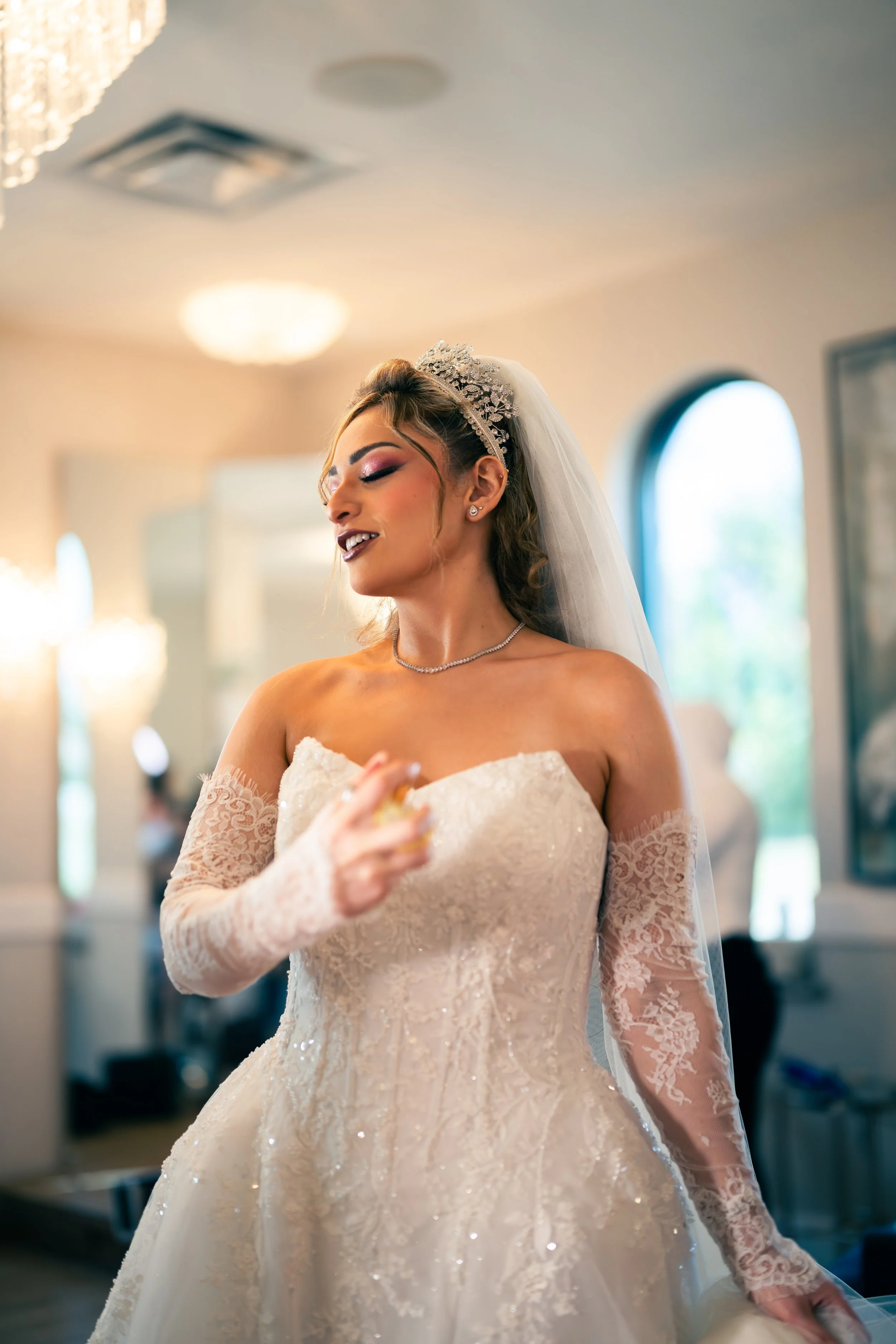 A bride in an elegant white wedding dress with lace sleeves, wearing a tiara and a veil, smiling and holding a perfume bottle in a warmly lit room.
