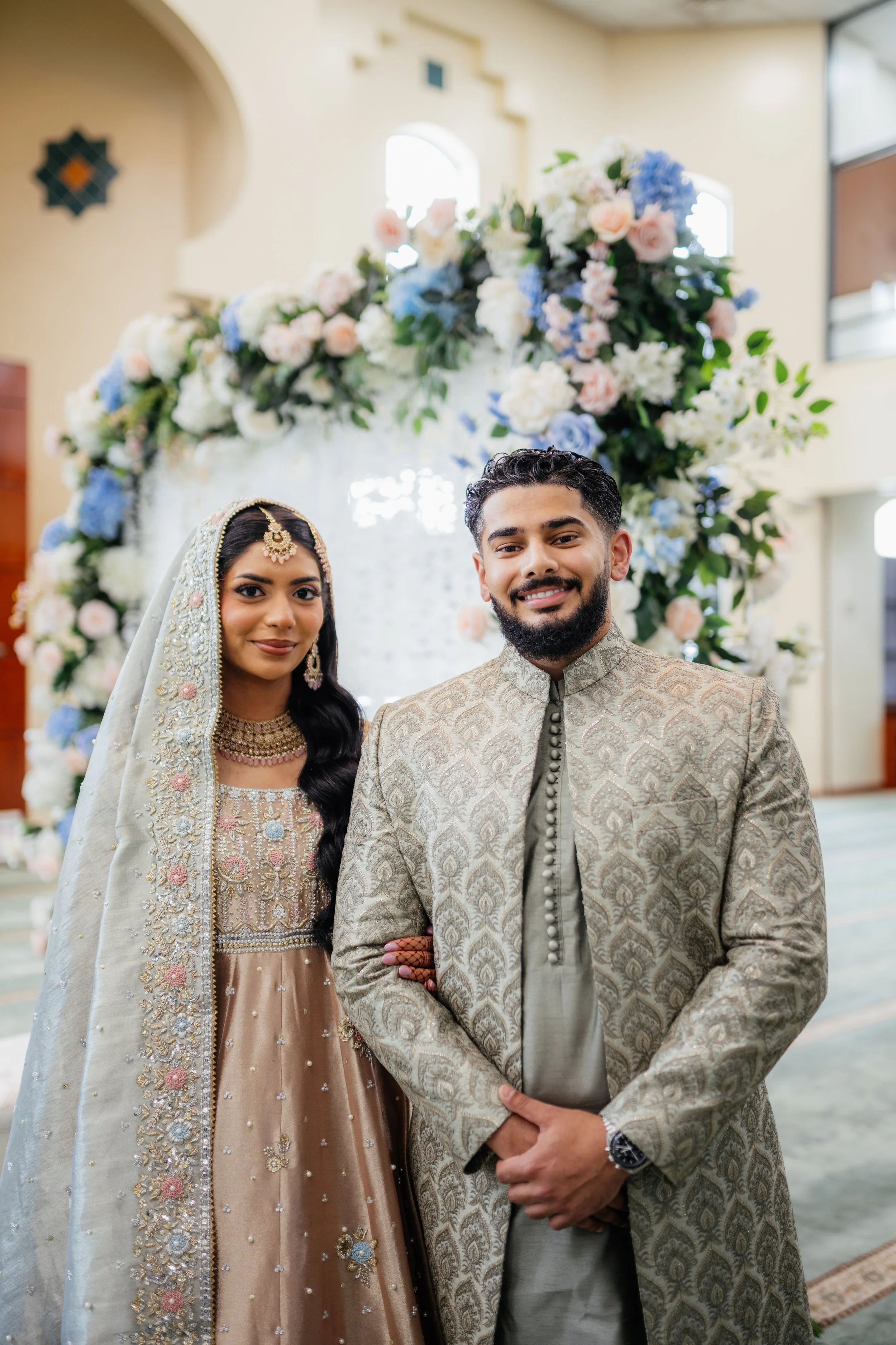 A bride and groom in traditional Indian wedding attire standing together in front of an elaborate floral backdrop.