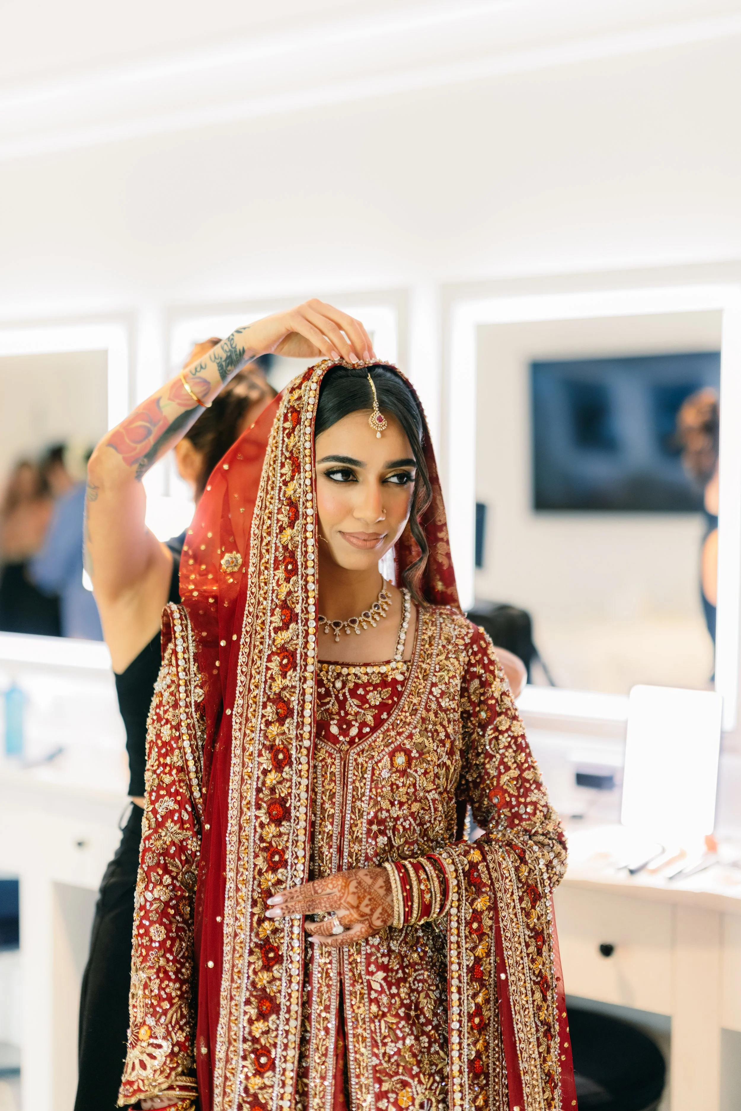 A woman dressed in traditional South Asian wedding attire, including a red and gold embroidered lehenga, ornate jewelry, and mehndi on her hands, is having her dupatta adjusted by another person in a bright, modern room.