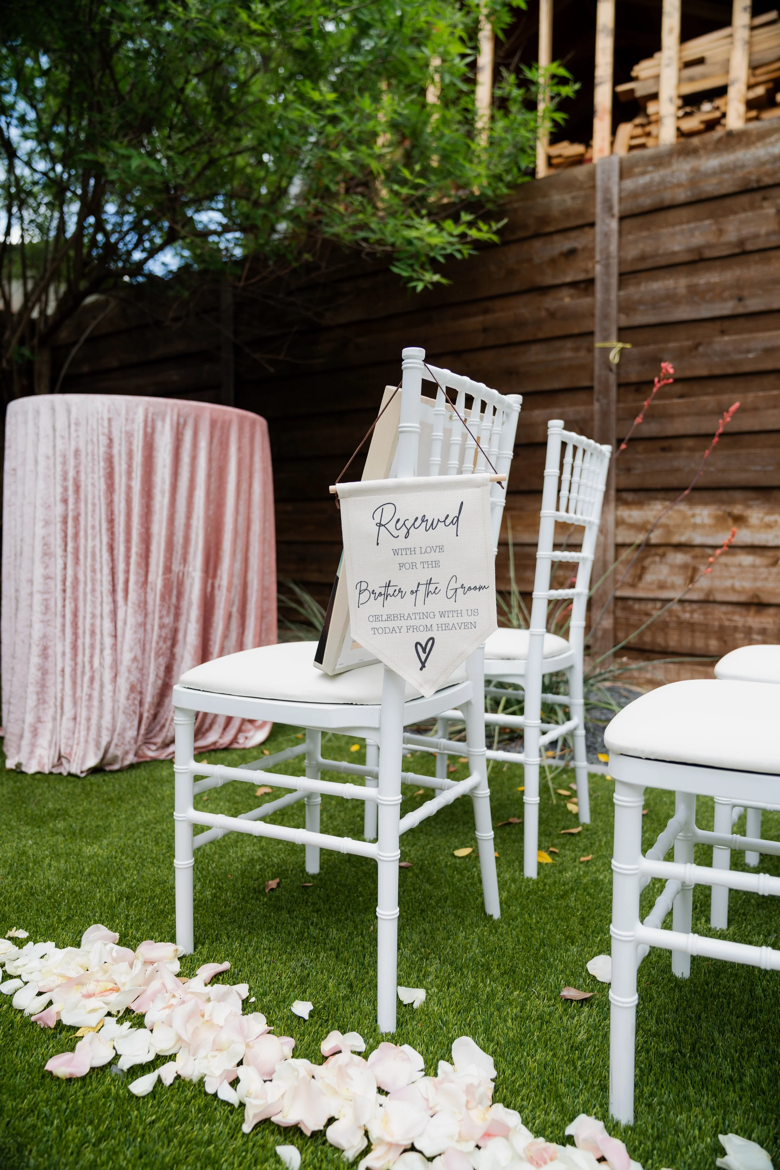 Wedding ceremony setup outdoors with white chairs, a table with a mauve tablecloth, and a reserved sign on one chair. Rose petals are scattered on the grass, and a wooden fence is visible in the background.