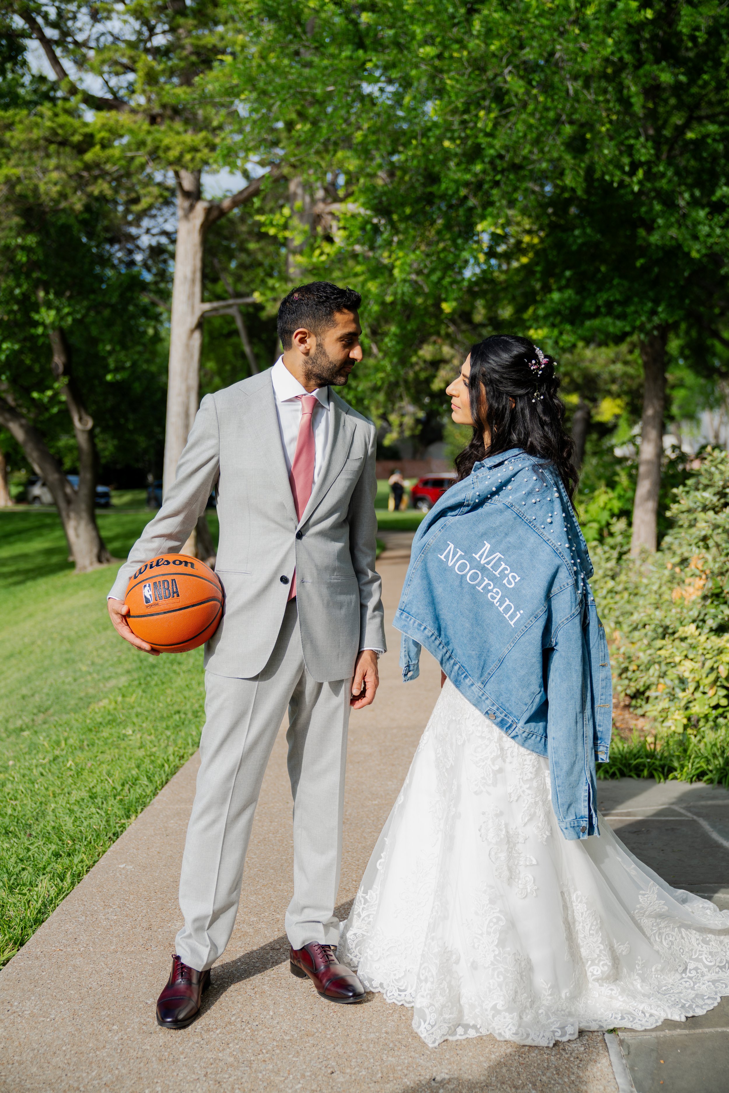 A man in a suit holding a basketball and a woman in a wedding dress with a denim jacket in a park, facing each other.