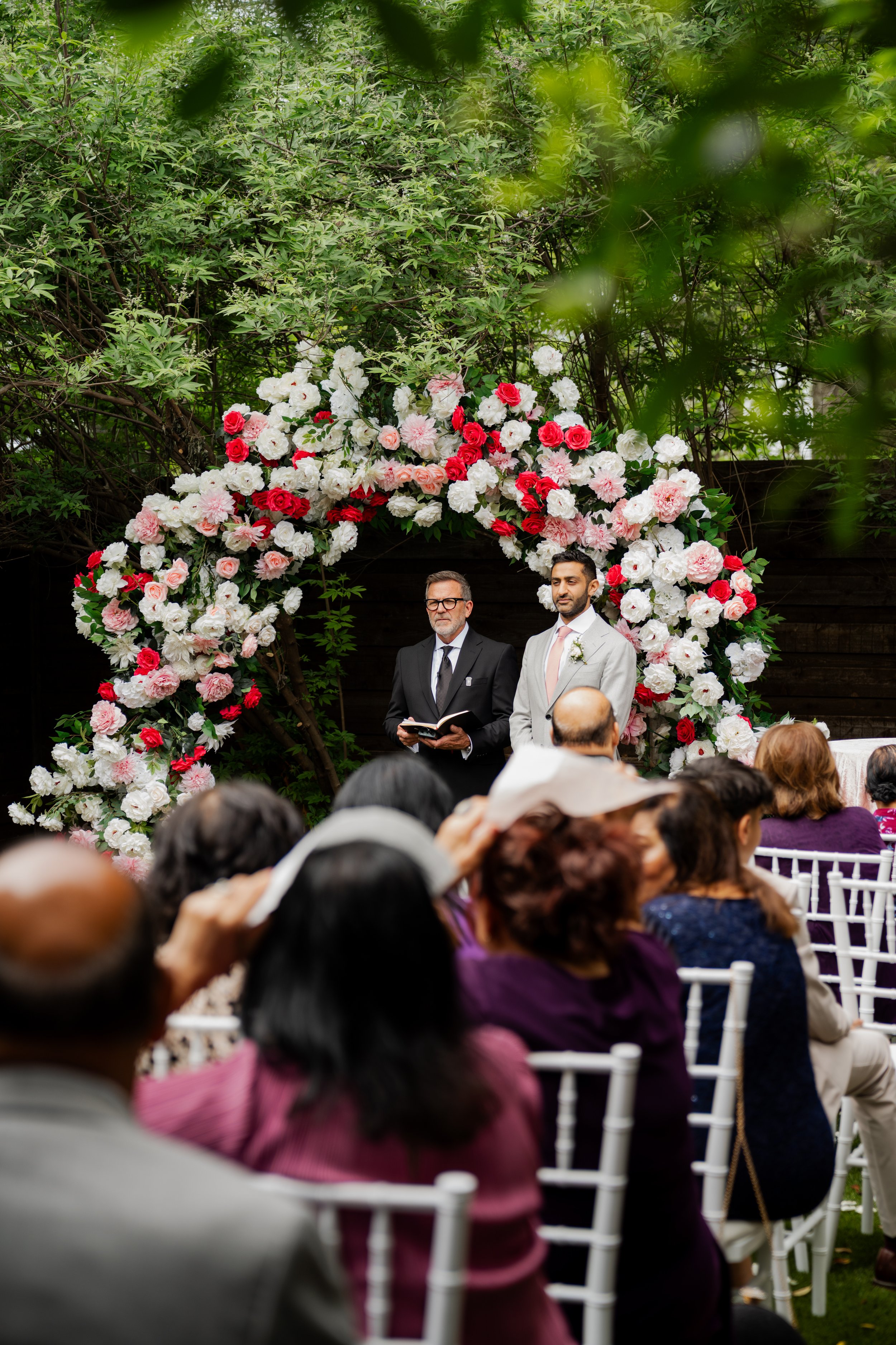 Two men standing under a large floral arch, one in a dark suit and glasses, the other in a light suit, during a wedding ceremony, surrounded by seated guests.