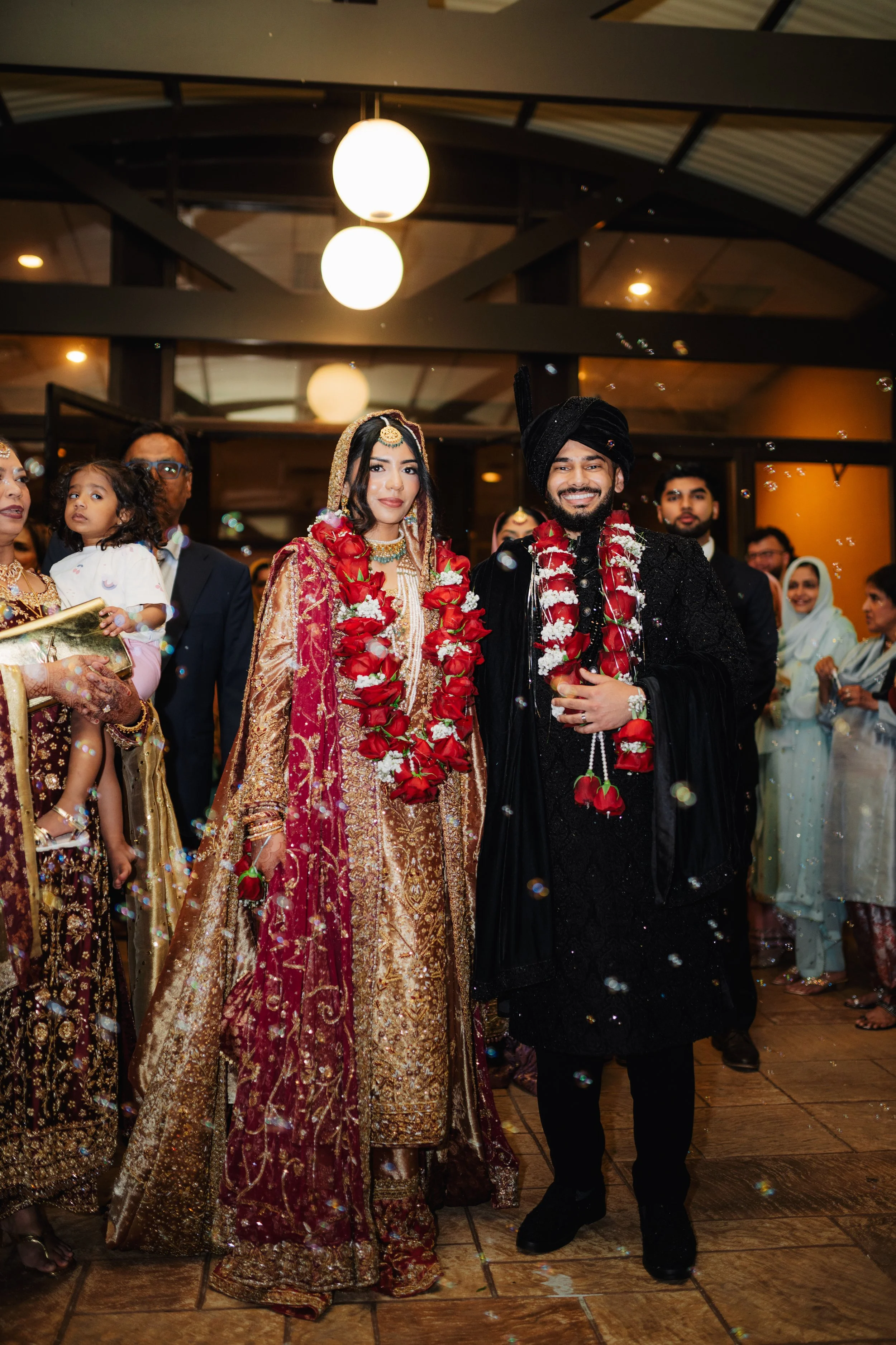 A bride and groom celebrate their wedding in traditional South Asian attire, adorned with flower garlands, surrounded by family and friends in an indoor venue with warm lighting.