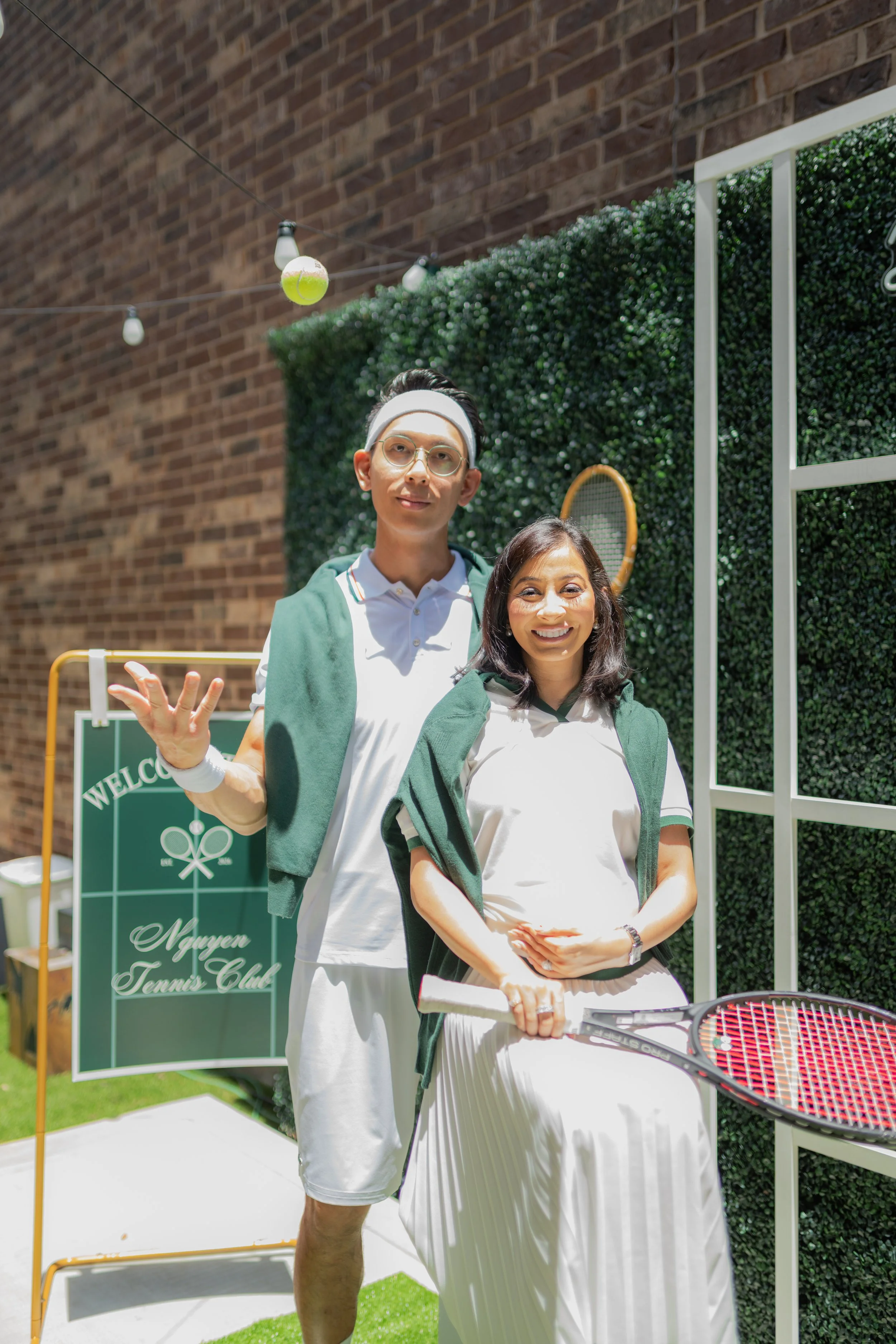 Two people, a man and a woman, standing outdoors on a tennis court with a Wimbledon tennis racket in front of them. The man is wearing glasses, a white tennis outfit, and a headband, and is waving. The woman is smiling, wearing a white dress and a gr