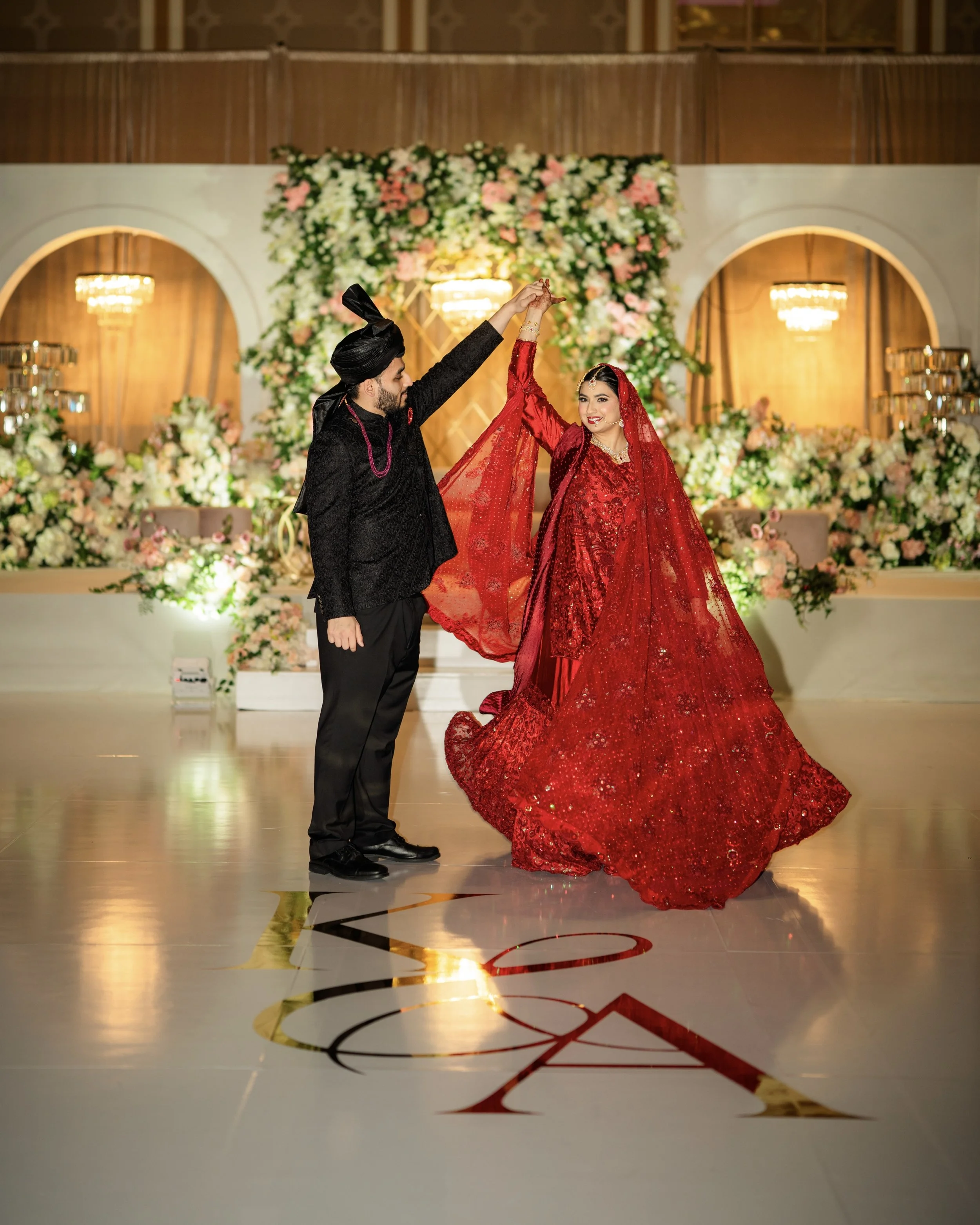 A bride and groom dancing at their wedding reception. The bride is wearing a red, heavily embroidered bridal gown with a matching red dupatta, and the groom is dressed in a black sherwani and turban. The background features a floral backdrop with pin